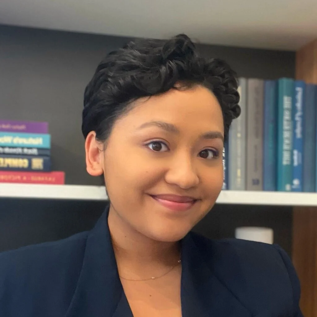 Close-up of a young woman with short, curly dark hair smiling in front of a bookshelf filled with books.