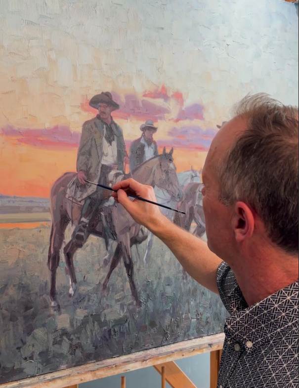 Man in blue striped collared shirt uses a small paint brush to add final touches to a western painting with two cowboys on horseback under a sun-setting sky.