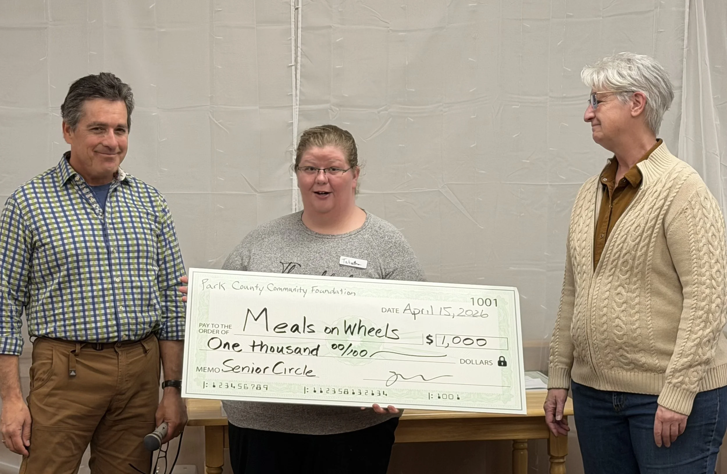 Man in plaid shirt gives large $1000 check to woman in grey sweater alongside a smiling woman with white hair and glasses.