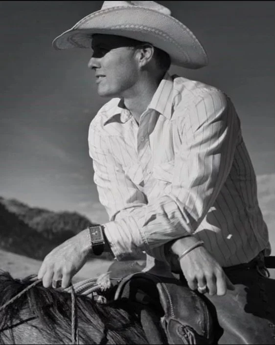 Black and white image of a middle-aged man on horseback wearing a white cowboy hat with a white shirt with his arms crossed and staring off into the distance to the left.