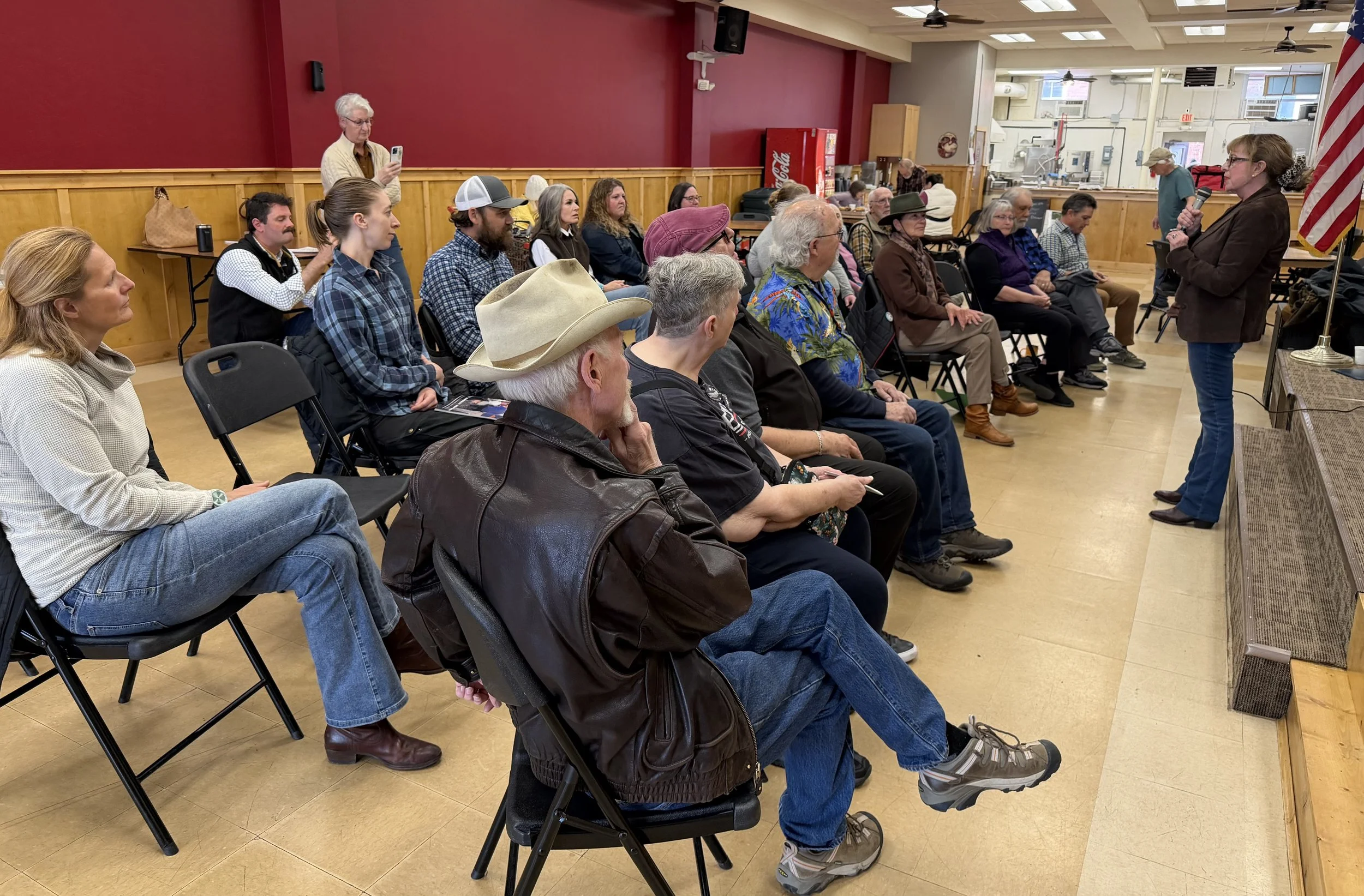 Woman with glasses, brown coat, and boots addresses a crowd of twenty adults and senior citizens in a large room.
