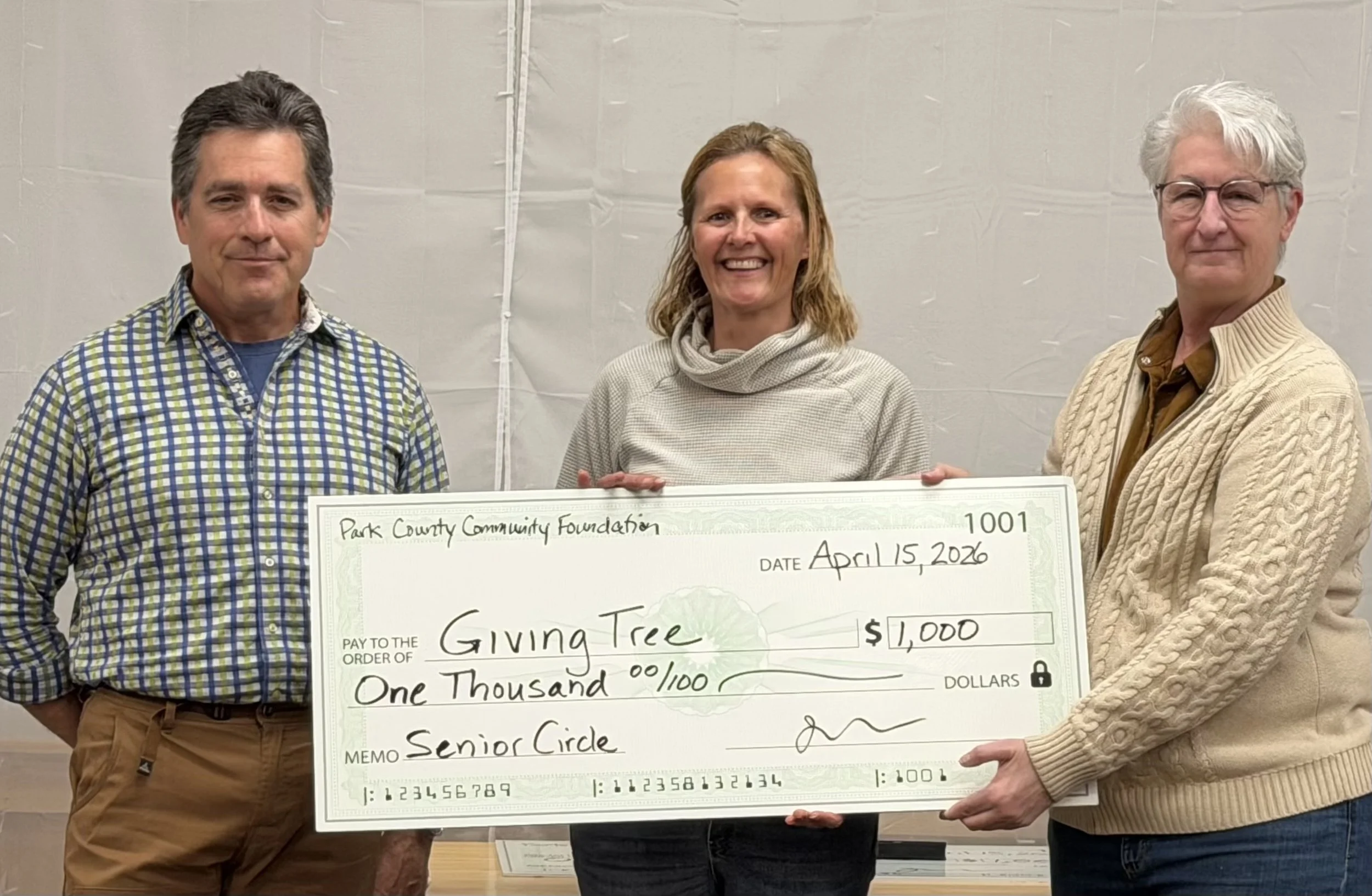 Man in plaid shirt presents large $1000 check to blonde woman in sweater next to a woman with white hair and glasses.