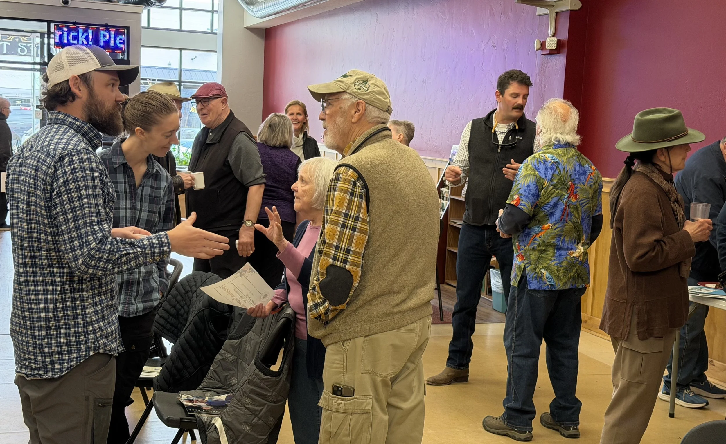Man in grey cap reaches out his hand for a handshake with an older man surrounded by people of various ages socializing and smiling.