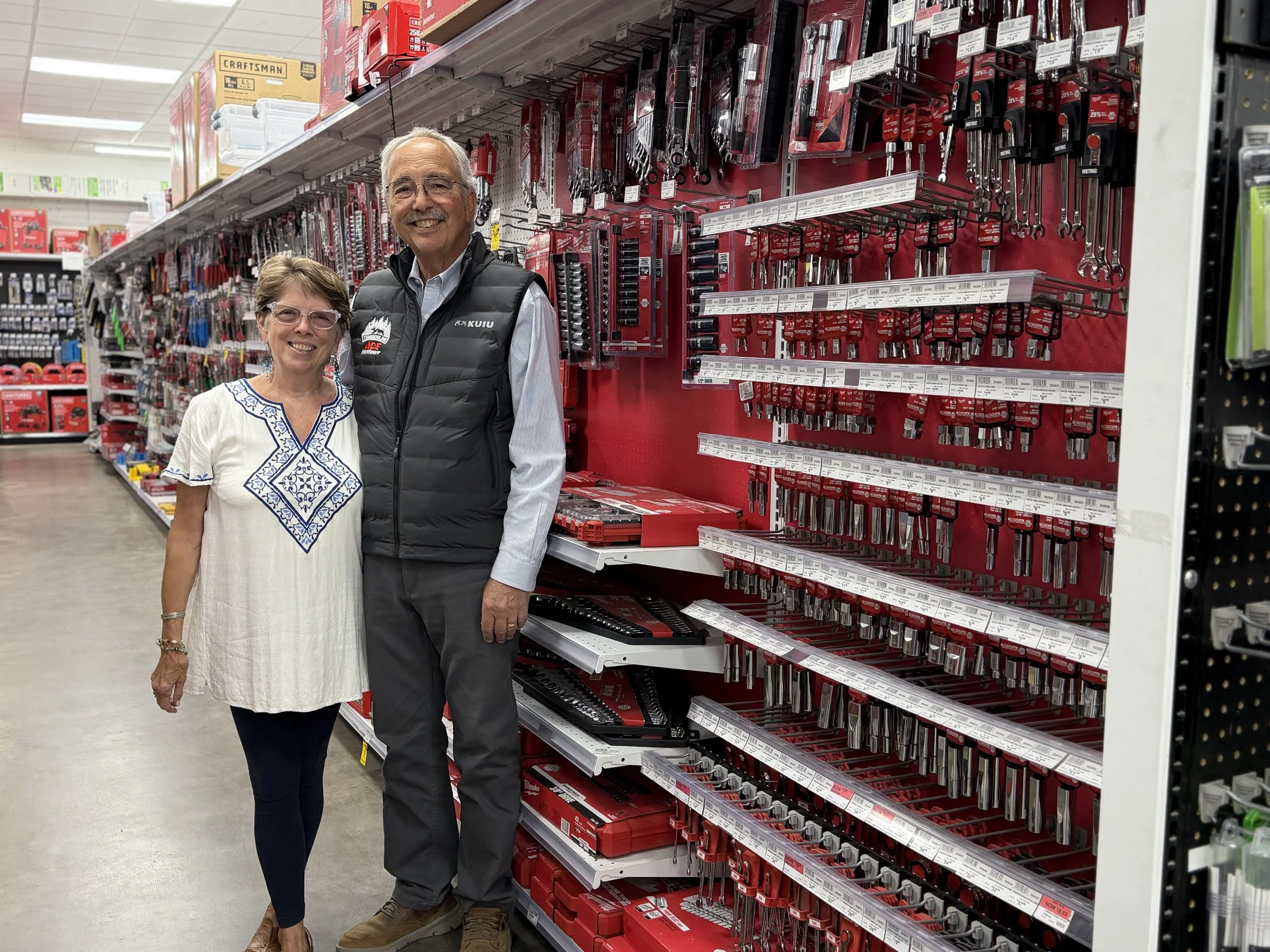 Woman in white blouse and glasses poses with a man in black vest while both are smiling and standing in the aisle of a tool store.