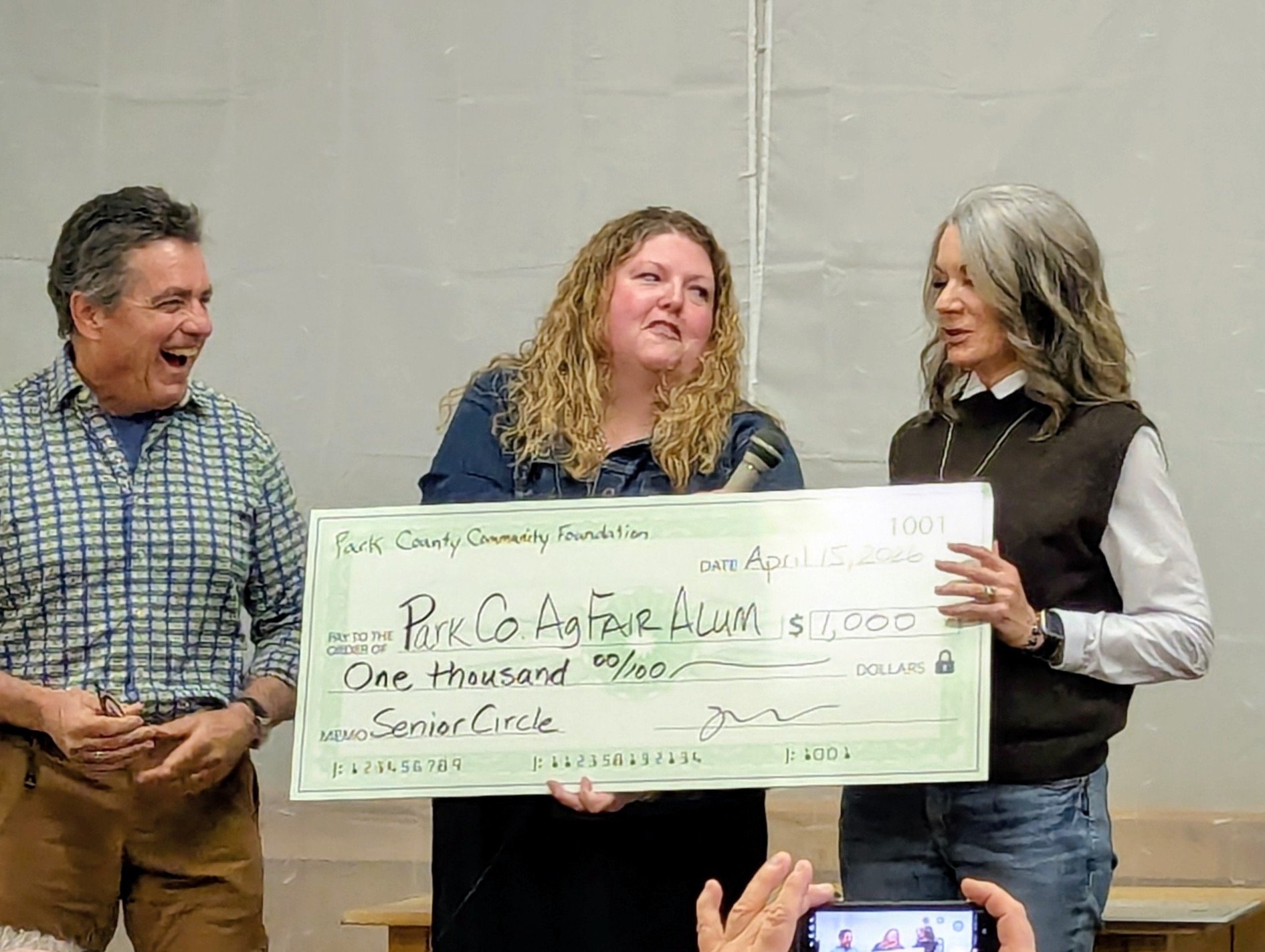 Happy man in plaid shirt presents large $1000 check to smiling blonde woman in denim top next to a woman with silver hair and brown vest.