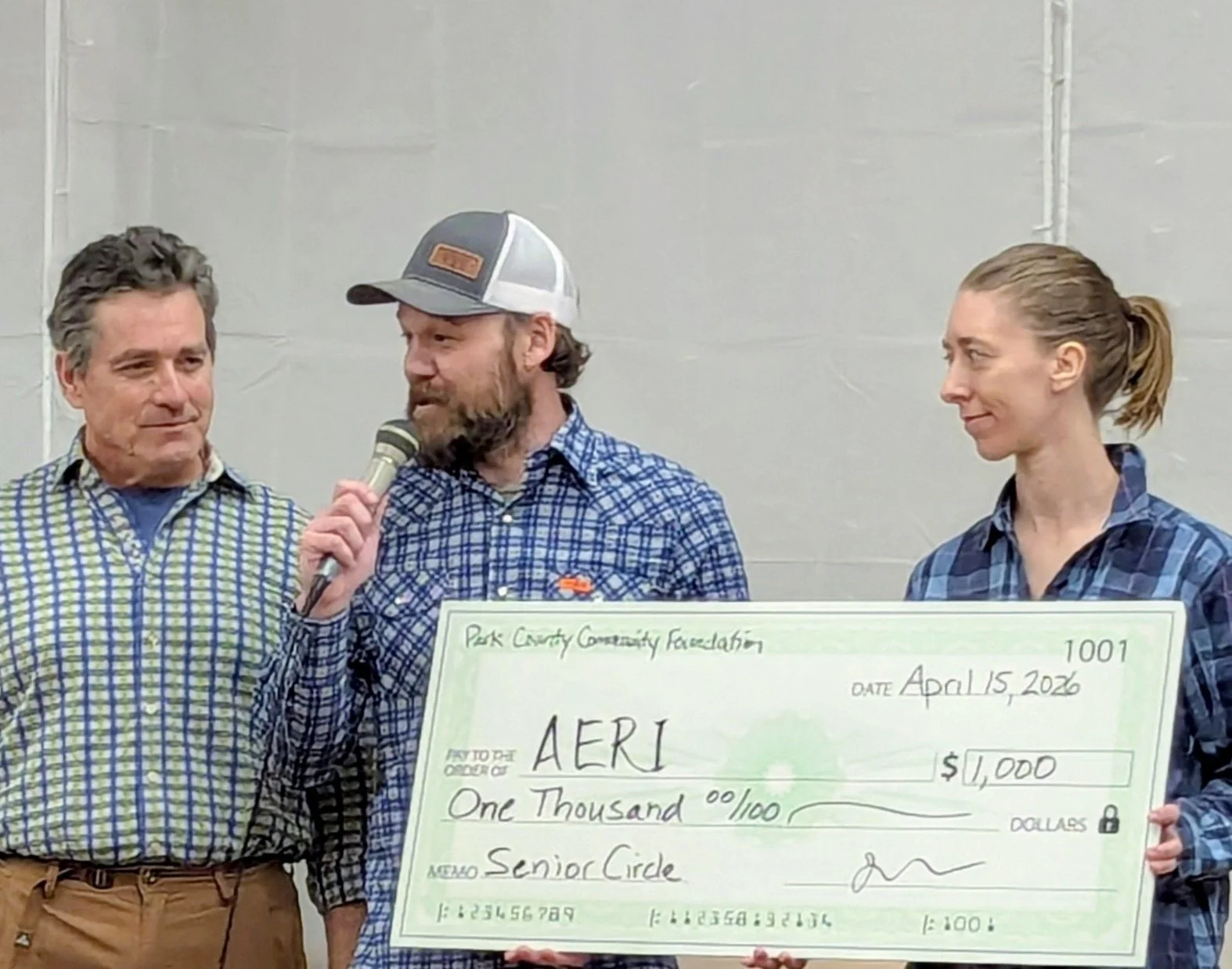 Man in plaid shirt presents large $1000 check to man in grey cap and woman with blonde ponytail