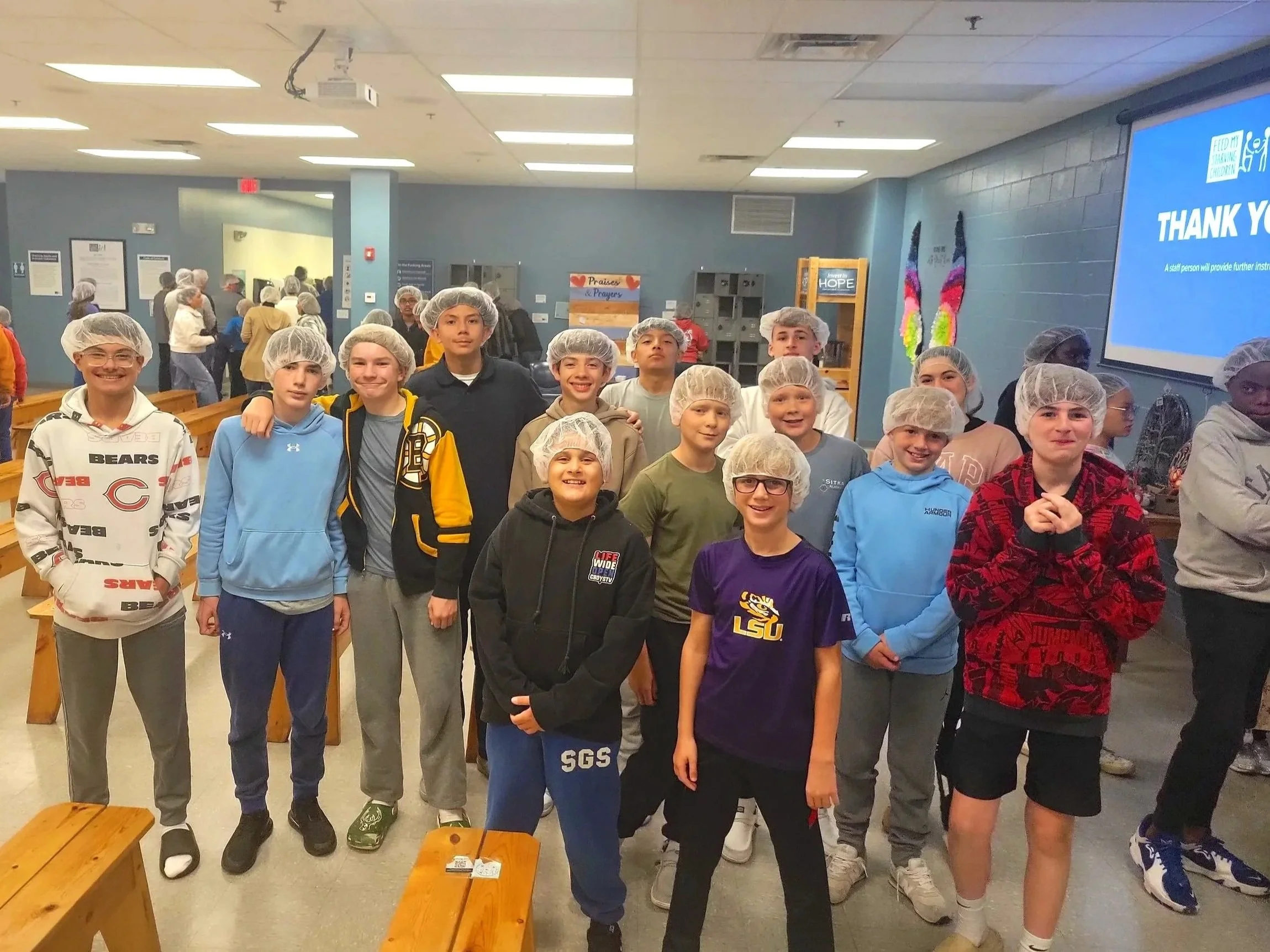 Group of children wearing hairnets inside a community room or cafeteria, some standing close together and smiling at the camera.