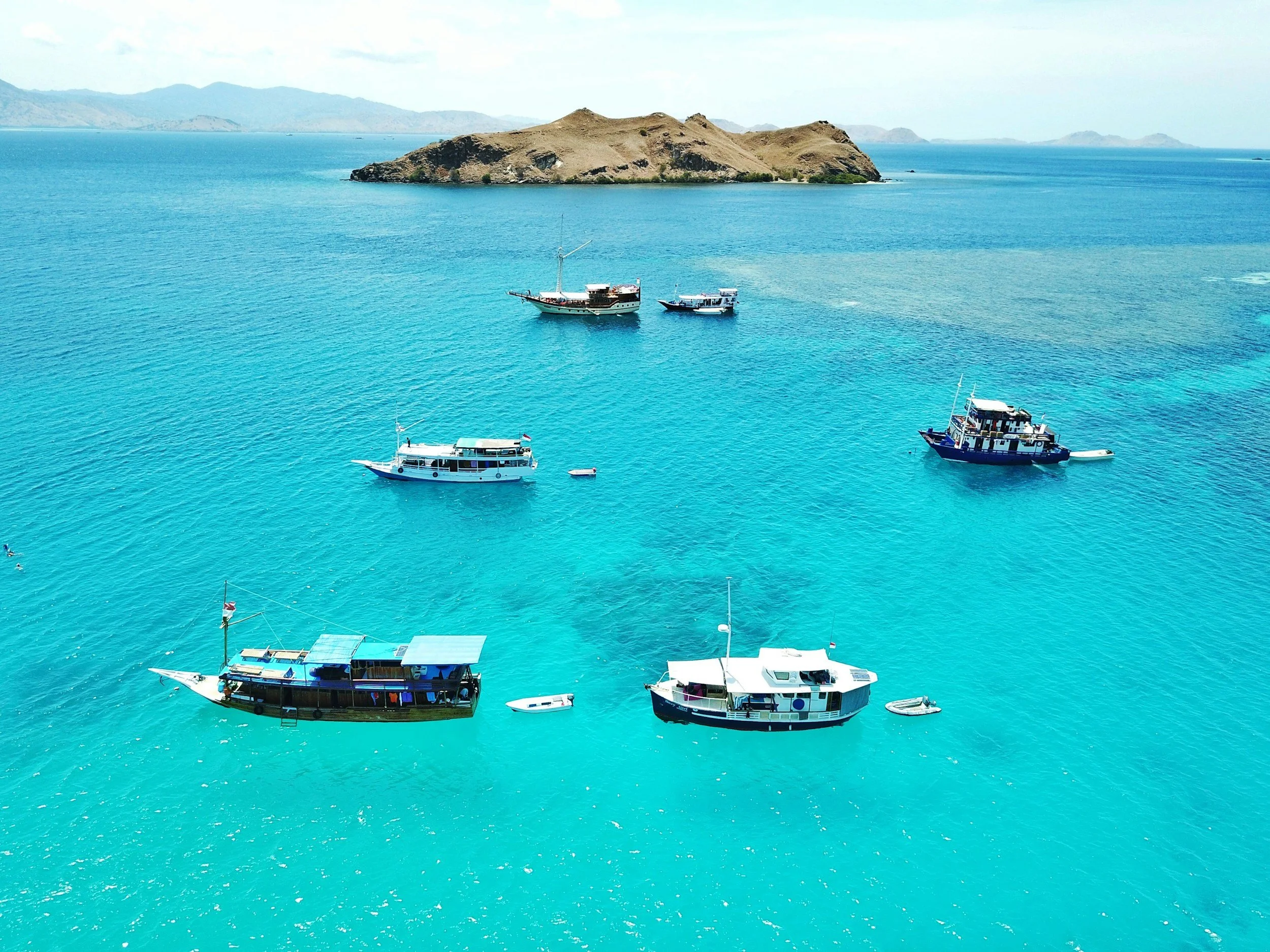 Several boats floating on clear turquoise water with an island and distant mountains in the background.