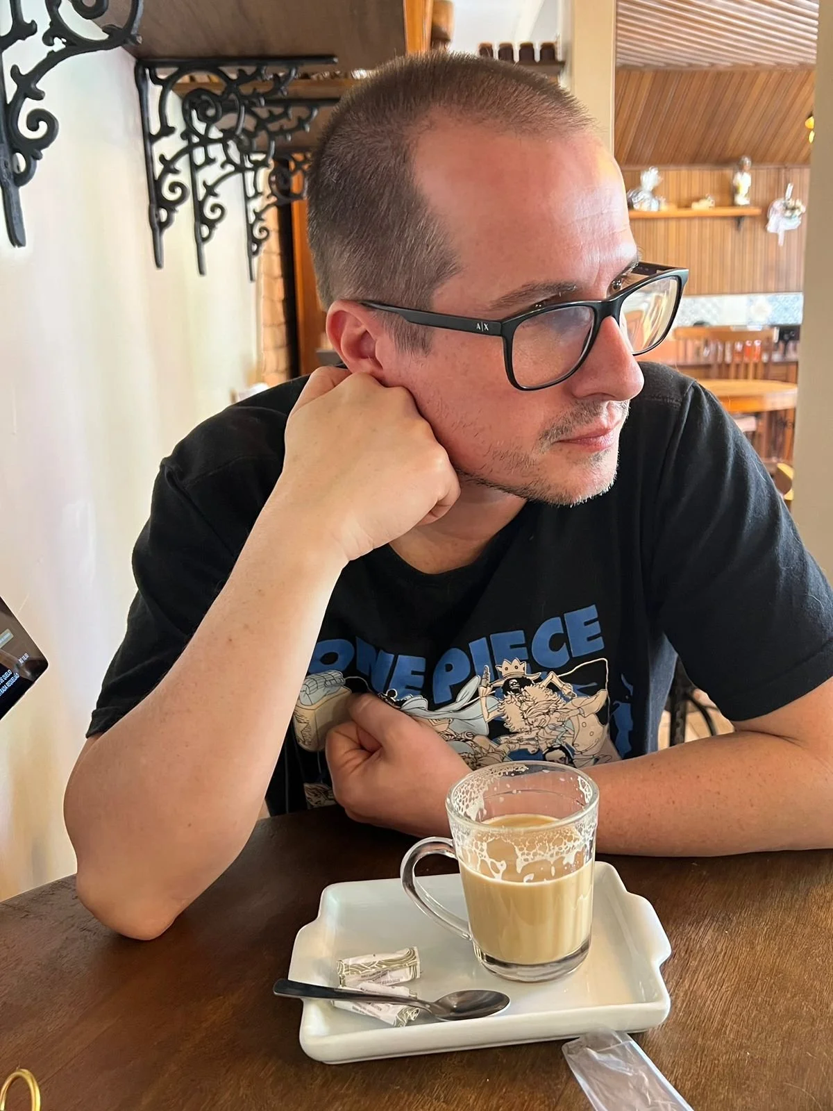 A man with glasses and a short haircut sitting at a table with a glass mug of coffee or tea, resting his chin on his hand, in a cozy restaurant or cafe with wooden interior decor.