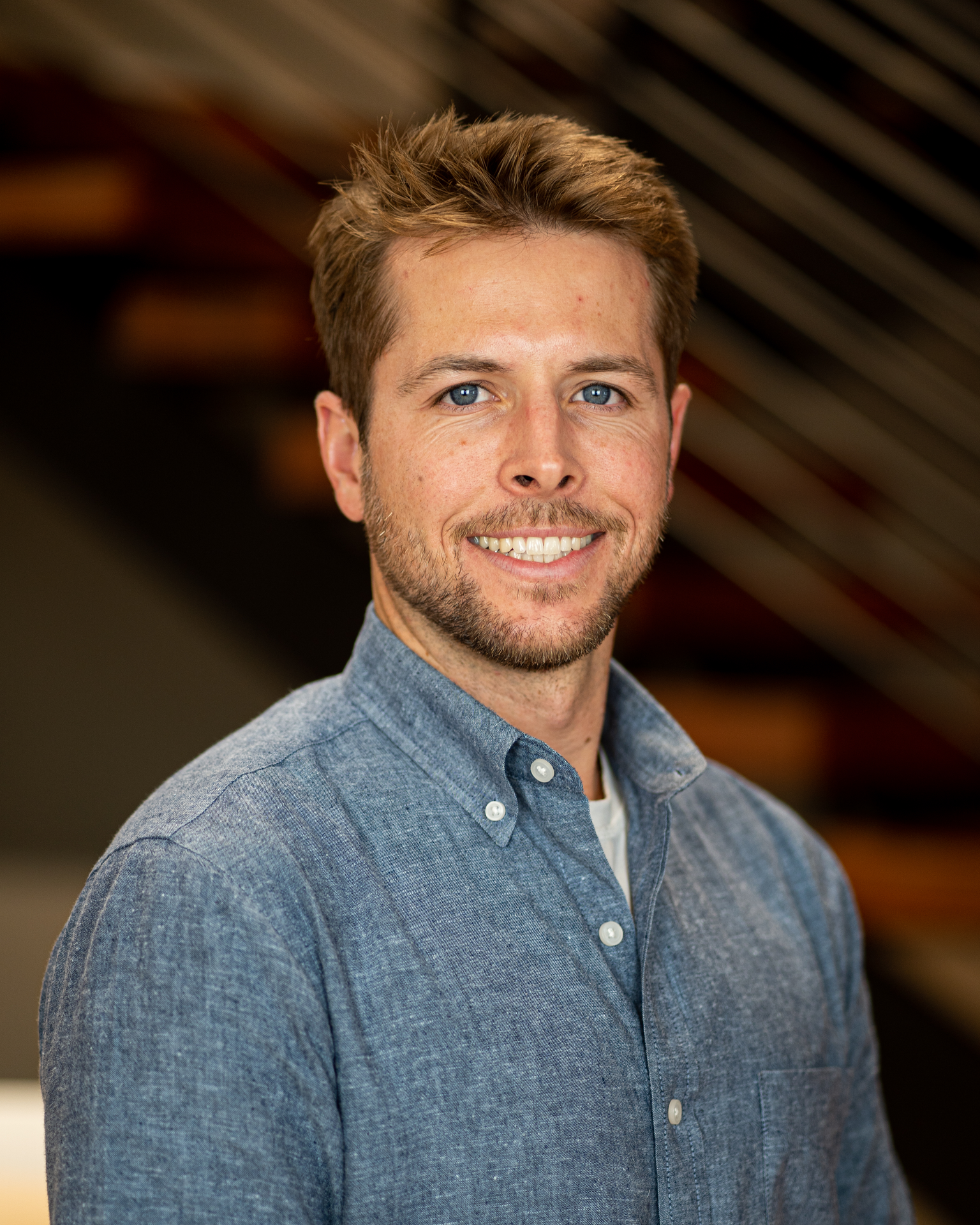 Smiling man in blue button-up shirt with blurred staircase in background.