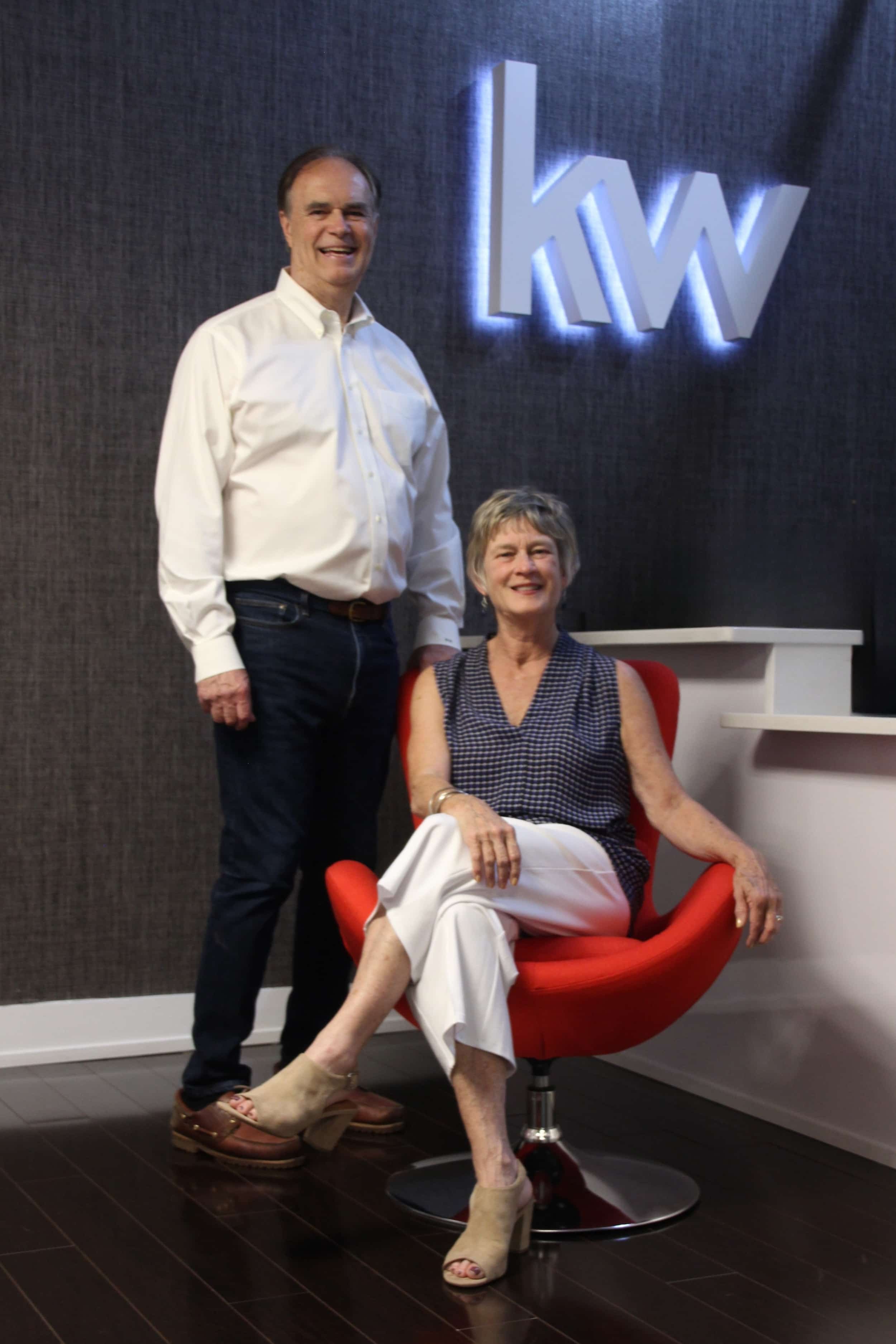 An older woman seated in a modern red chair with a round base, wearing a sleeveless black and white dress, beige open-toed high heels, and white pants, smiling at the camera. A man stands beside her, wearing a white button-up shirt, dark jeans, and brown sandals, also smiling. They are in a room with a dark textured wall and a backlit logo with the letters 'KW' on the wall.