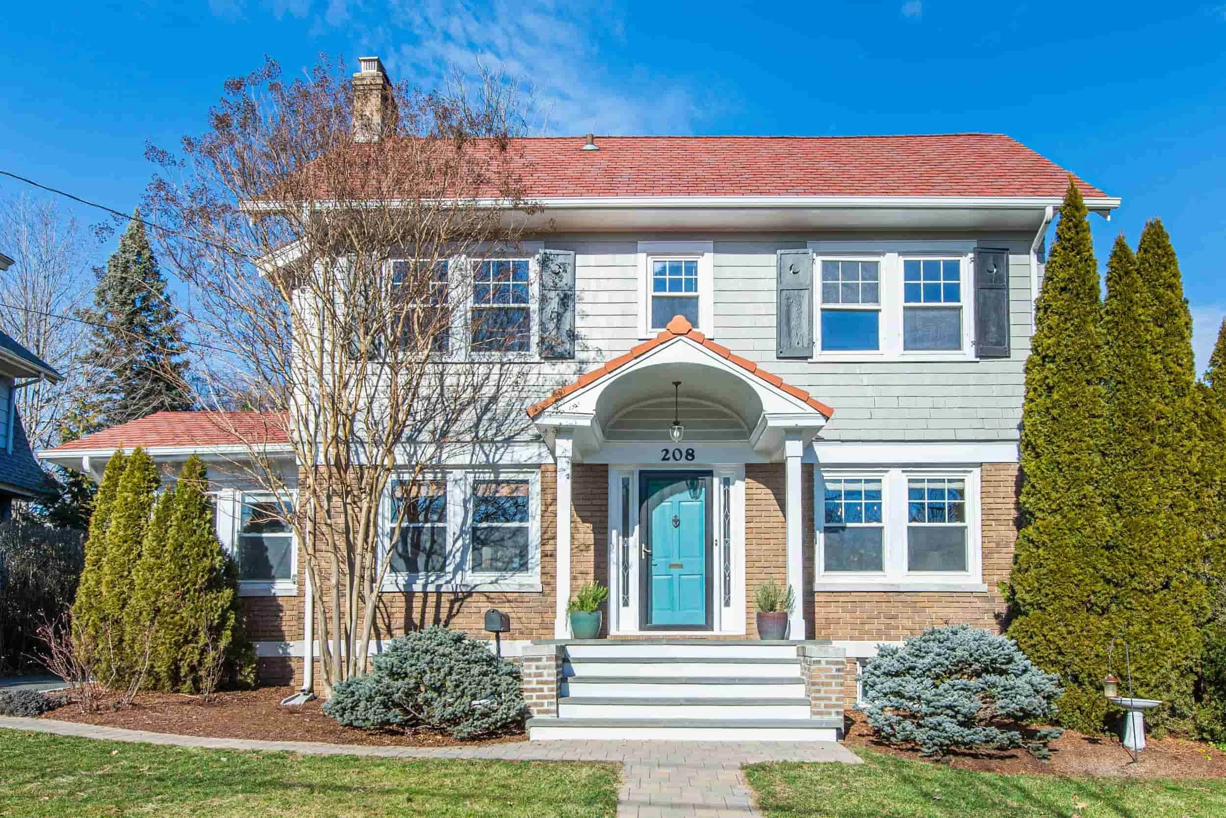A two-story house with a light blue front door, brick and gray siding, and multiple windows. There are trees and bushes in the yard, with a clear blue sky above.