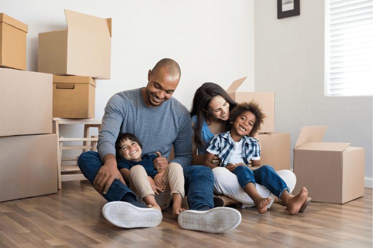 Family of four sitting on the floor among moving boxes in a new home, smiling and enjoying each other's company.