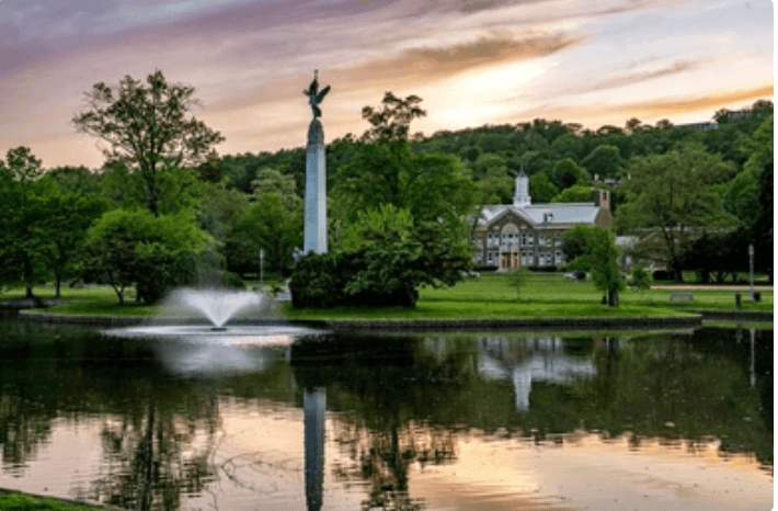 A park scene with a body of water reflecting trees, a fountain, and a monument, with a building in the background during sunset.