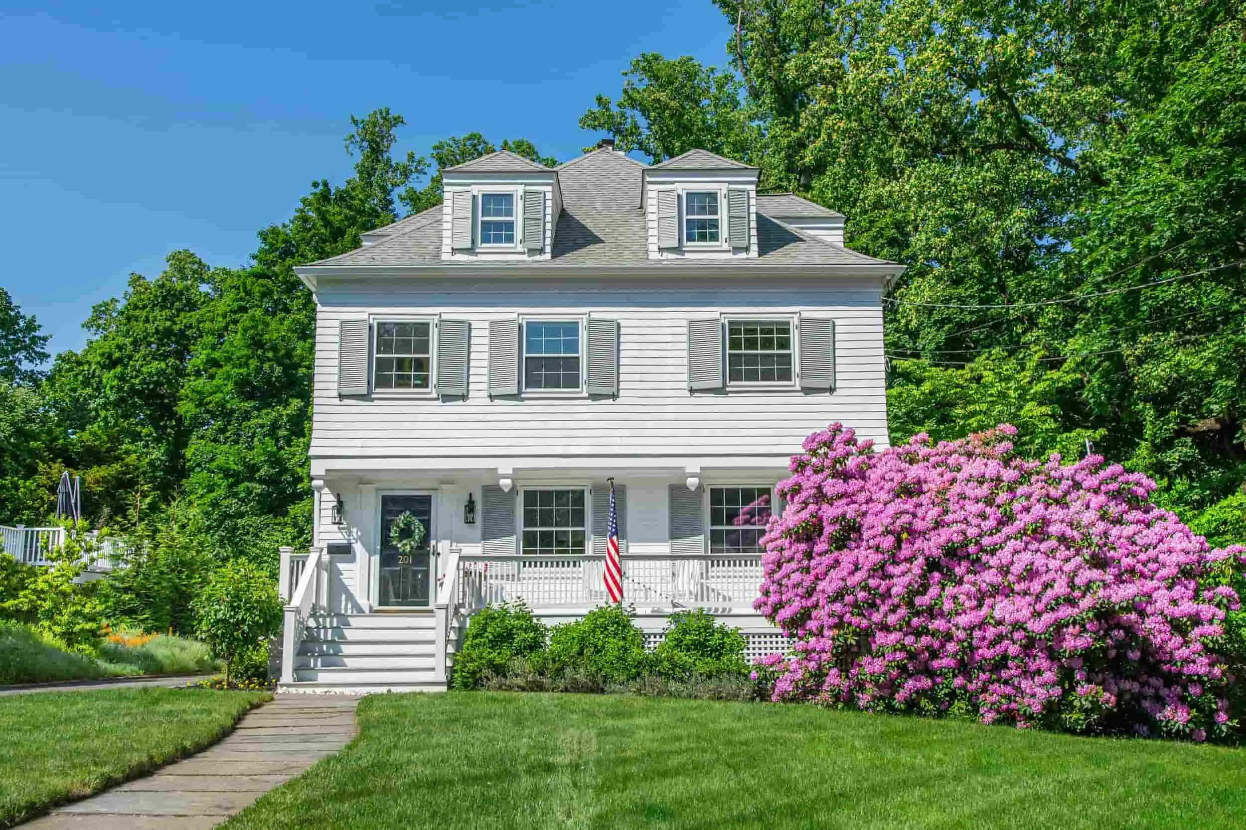 White house with front porch, surrounded by green grass and pink flowering bushes, with a blue sky and tall trees in the background.
