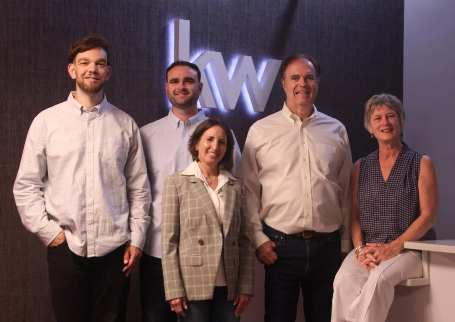 Group of six people standing together in front of a wall with a lit-up logo. They are smiling and dressed in business casual attire.