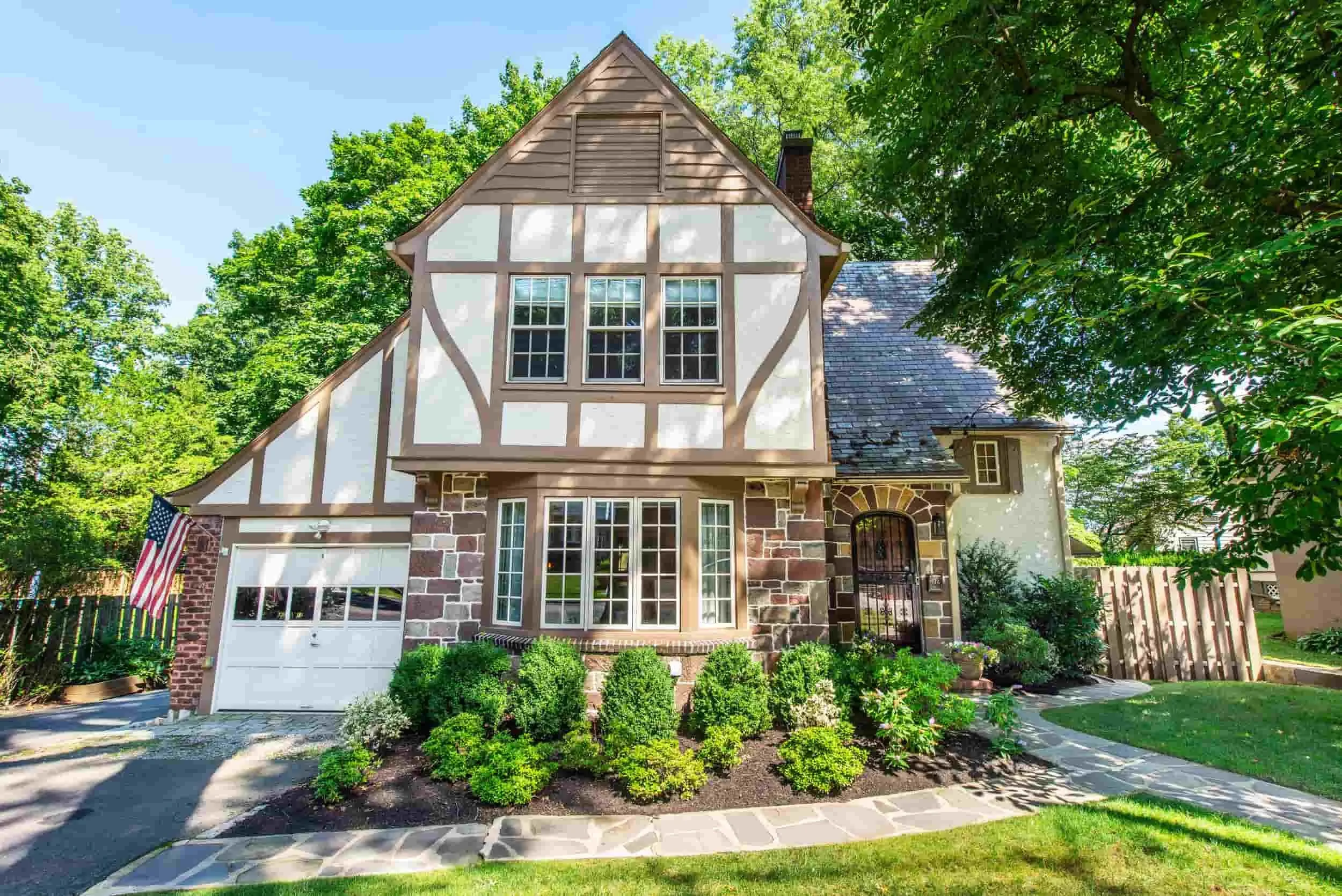 A two-story house with a Tudor-style facade, brick and stone lower level, and white upper level with wooden beams and large windows. The front yard has green shrubbery and a paved walkway, surrounded by tall trees and a wooden fence.