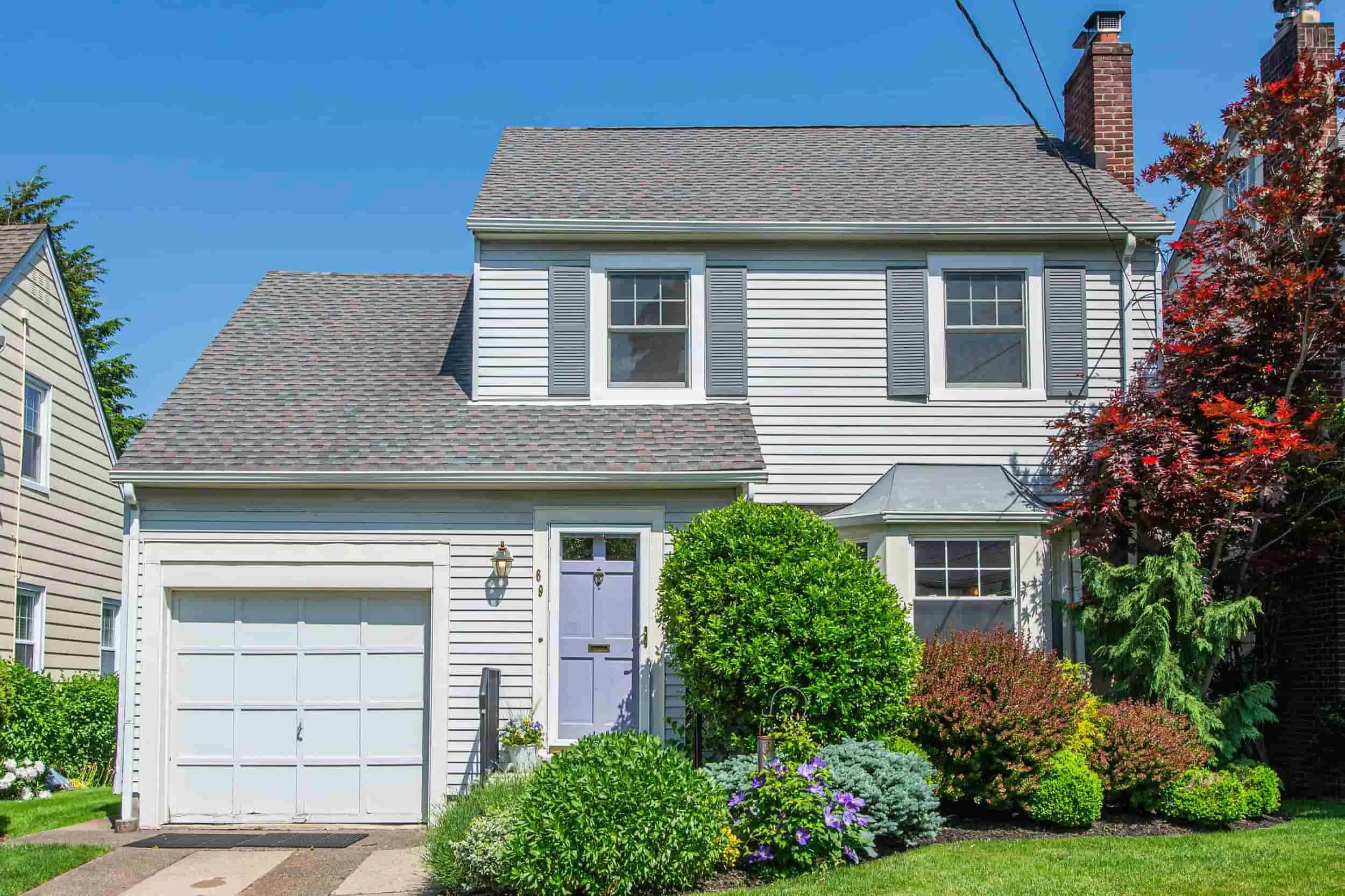 Front view of a two-story house with white siding, gray shutters, a purple front door, and a white garage door, surrounded by green shrubs, colorful flowers, and trees, with a clear blue sky overhead.