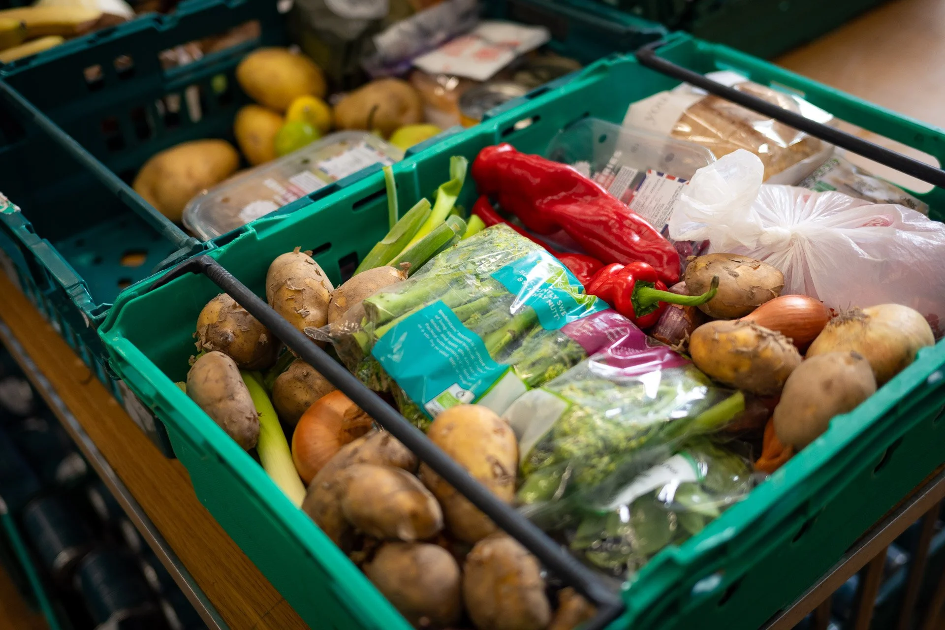 An OMA food parcel - a Green plastic crate filled with potatoes, green onions, red bell peppers, onions, garlic, and other vegetables, on a wooden table.