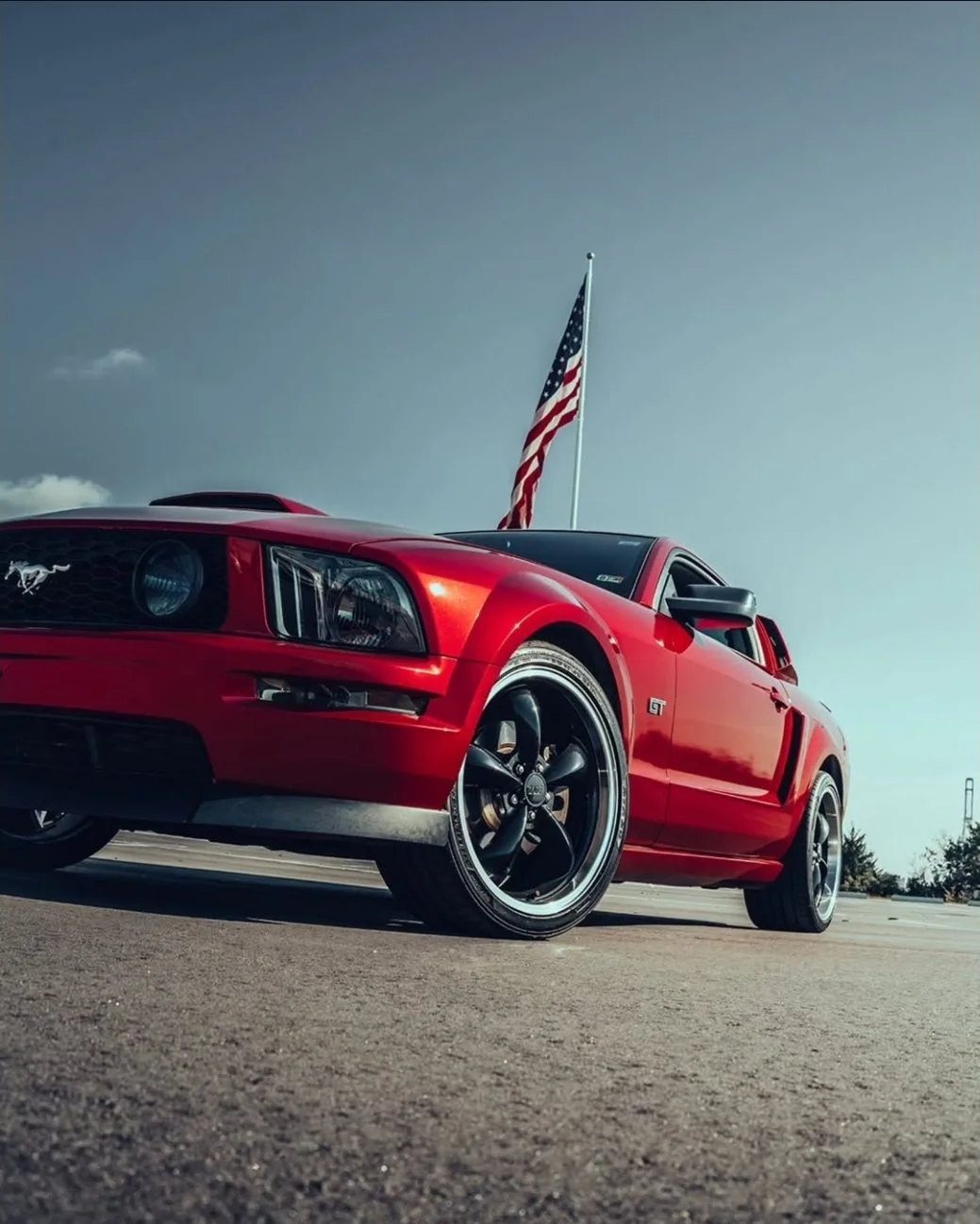 Red Ford Mustang GT with black wheels parked outdoors, American flag flying behind it against blue sky.