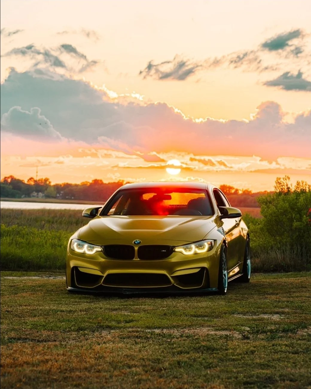 A yellow BMW car parked on grass with a sunset sky, clouds, and a body of water in the background.