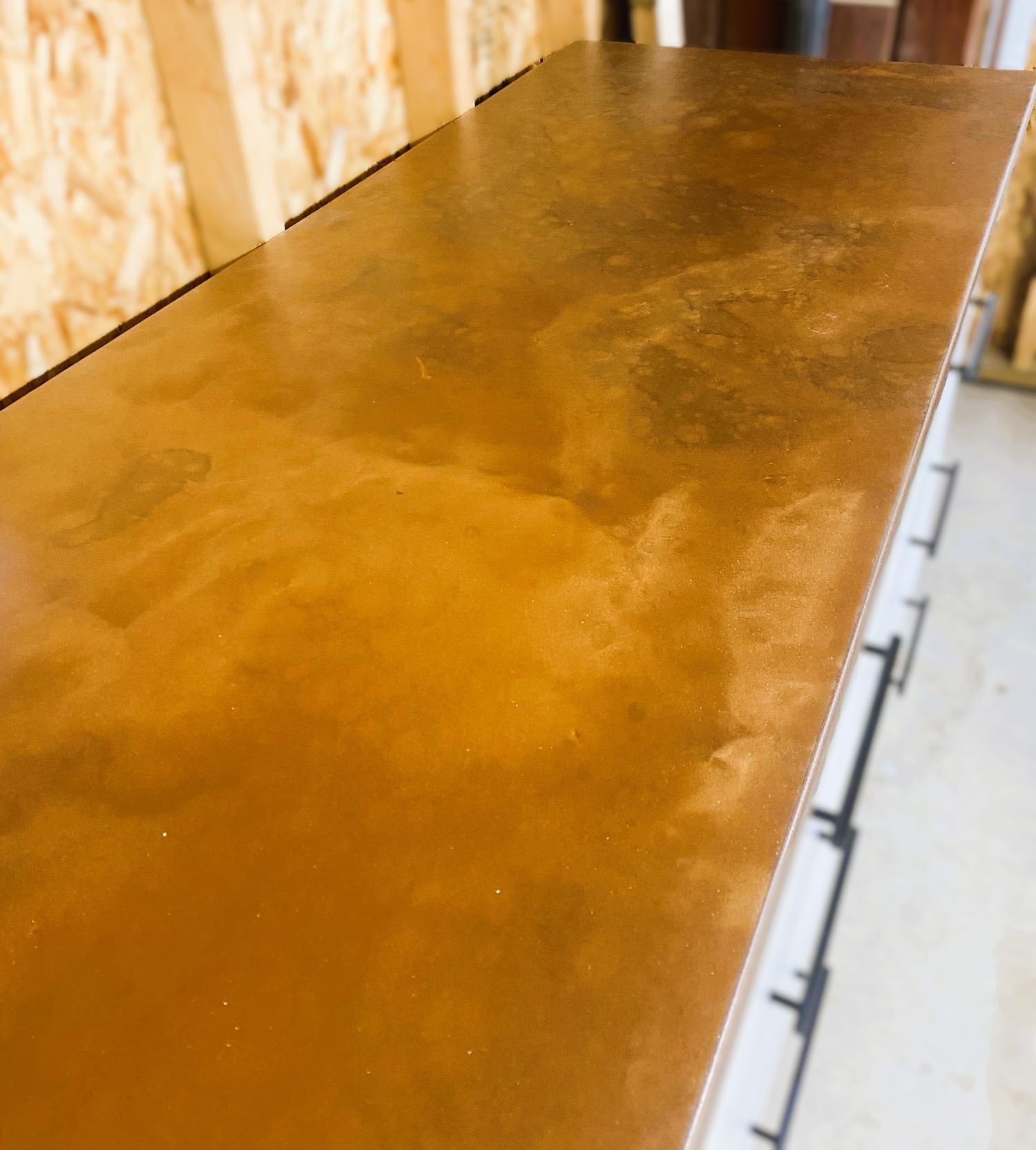 A close-up view of a wooden countertop or tabletop with a polished, glossy finish. The background shows a wall with unfinished wood paneling and some black handles.