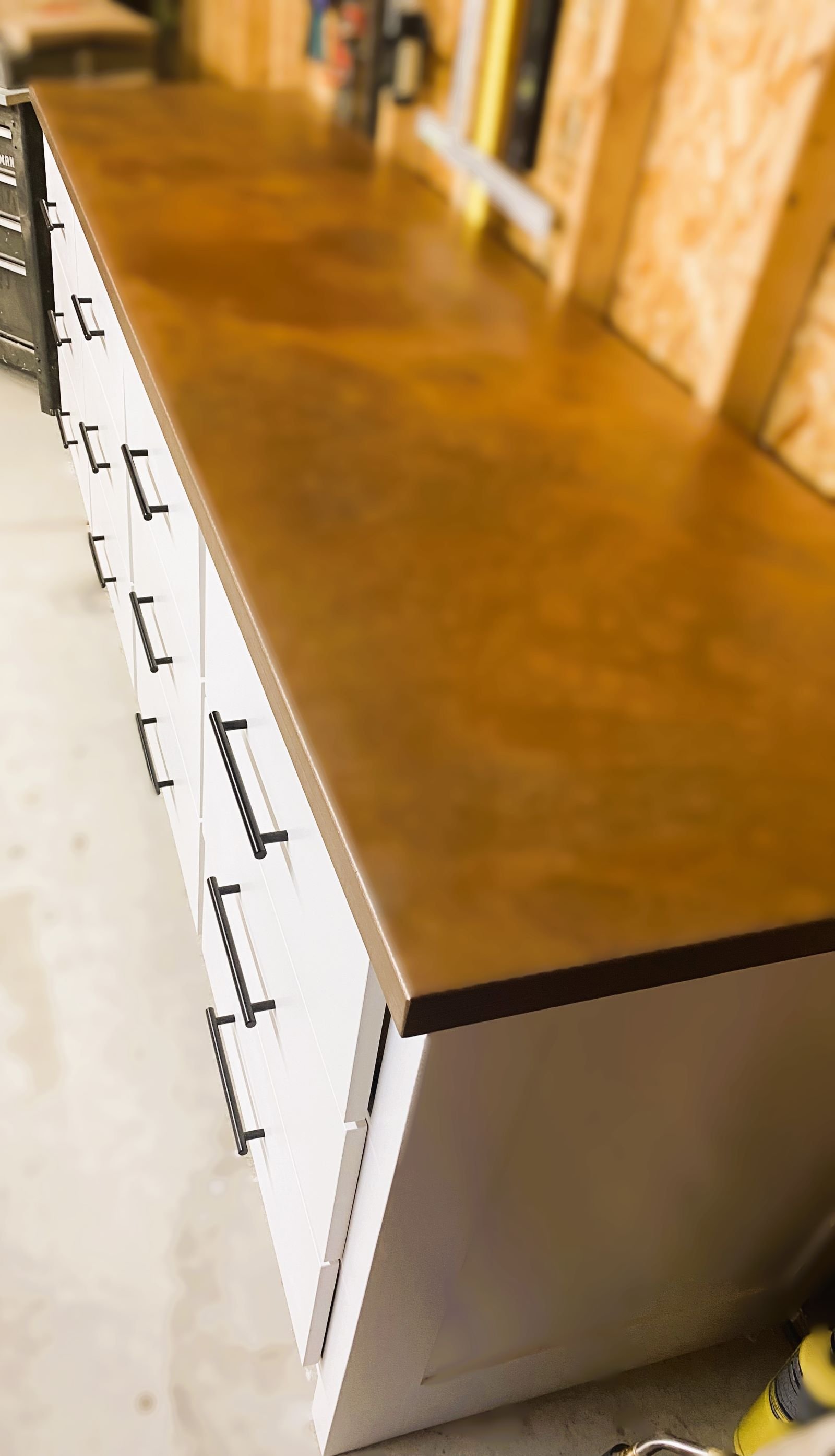 Close-up of a wooden countertop with white cabinets and black handles below. The background shows a wood-paneled wall with shelves.