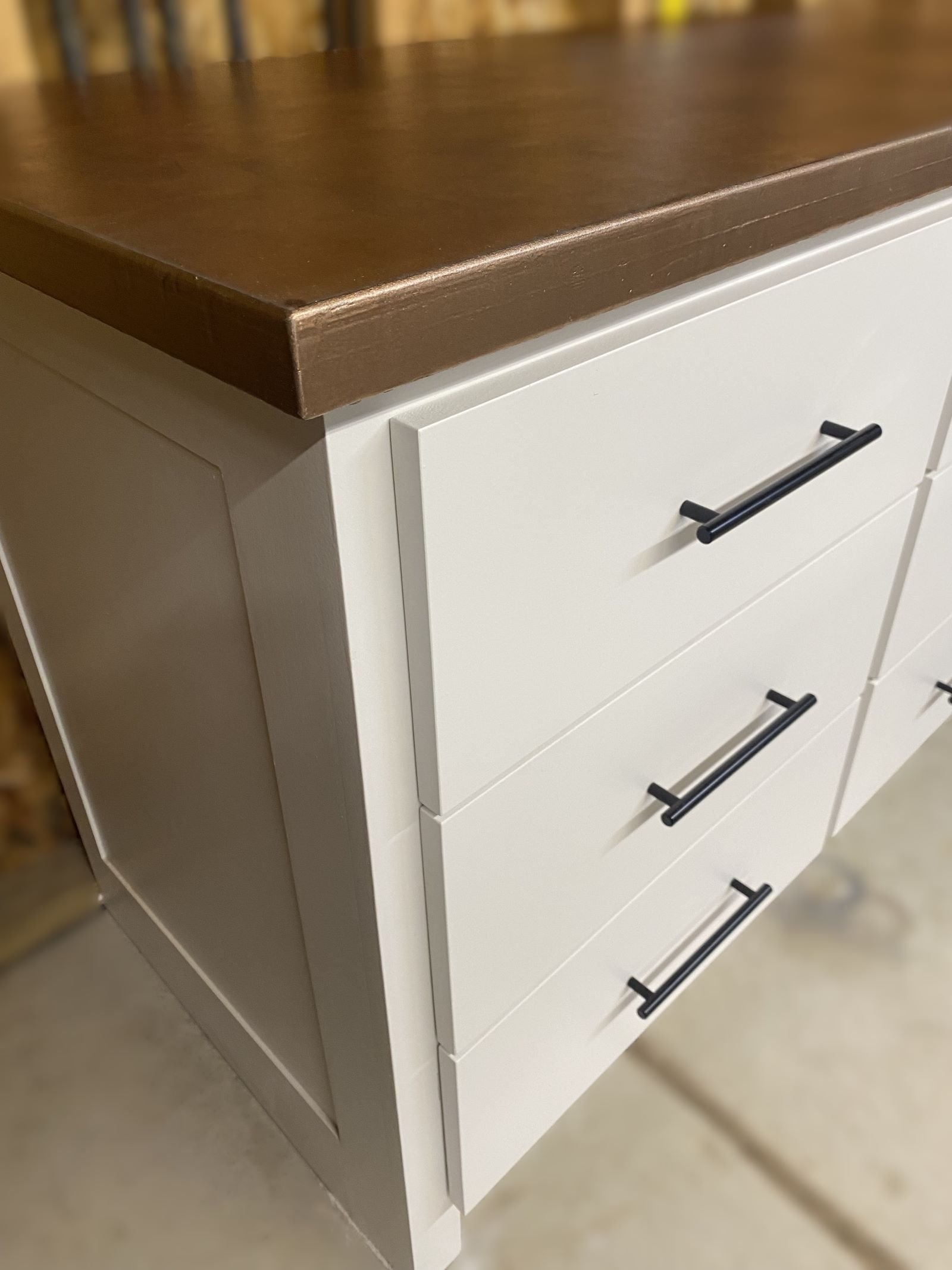 Close-up of a white dresser with three drawers and black handles, topped with a wooden surface.
