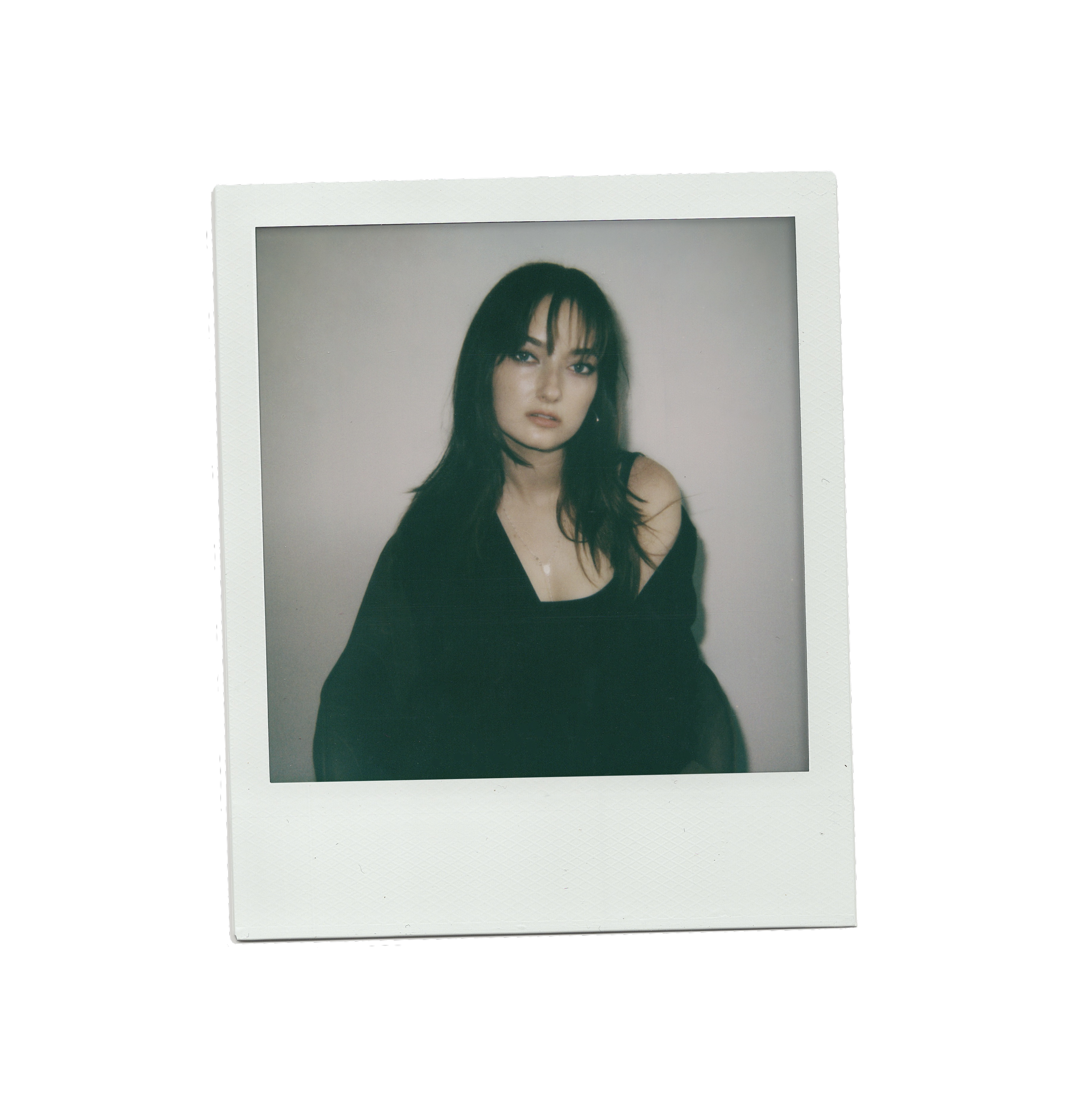 A woman with dark hair and bangs, wearing a black off-the-shoulder top, looking at the camera with a neutral expression, against a plain light-colored wall.