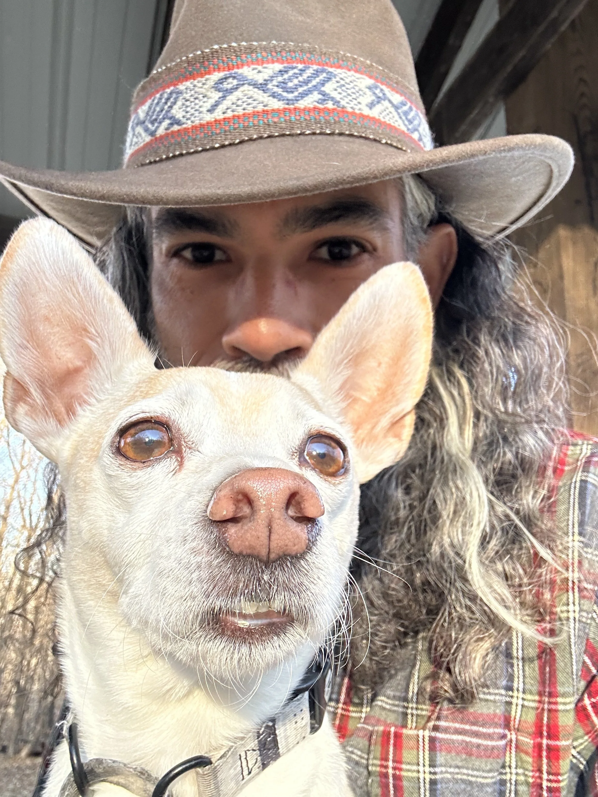 A person wearing a brown cowboy hat with a decorative band, taking a close-up selfie with a tan-colored dog with large ears, amber eyes, and a black collar.