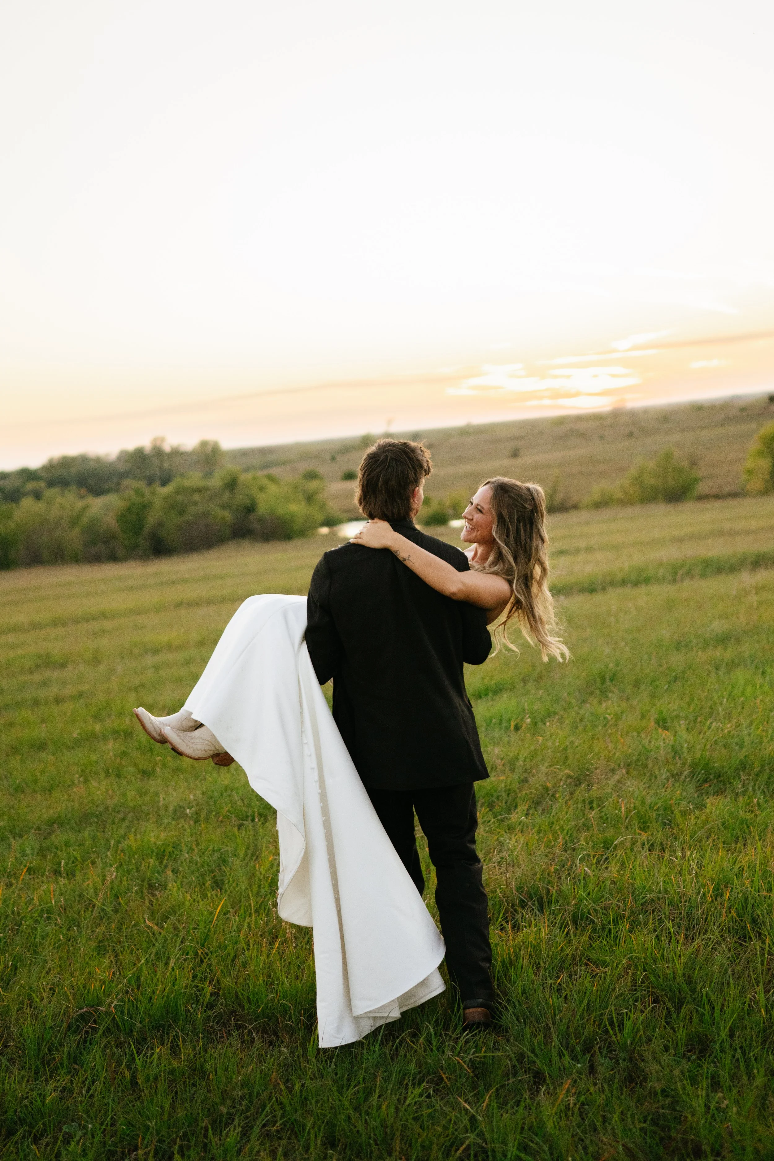 A couple, with the woman in a white wedding dress and the man in a black suit, enjoying a sunset in a grassy field. The man is carrying the woman.