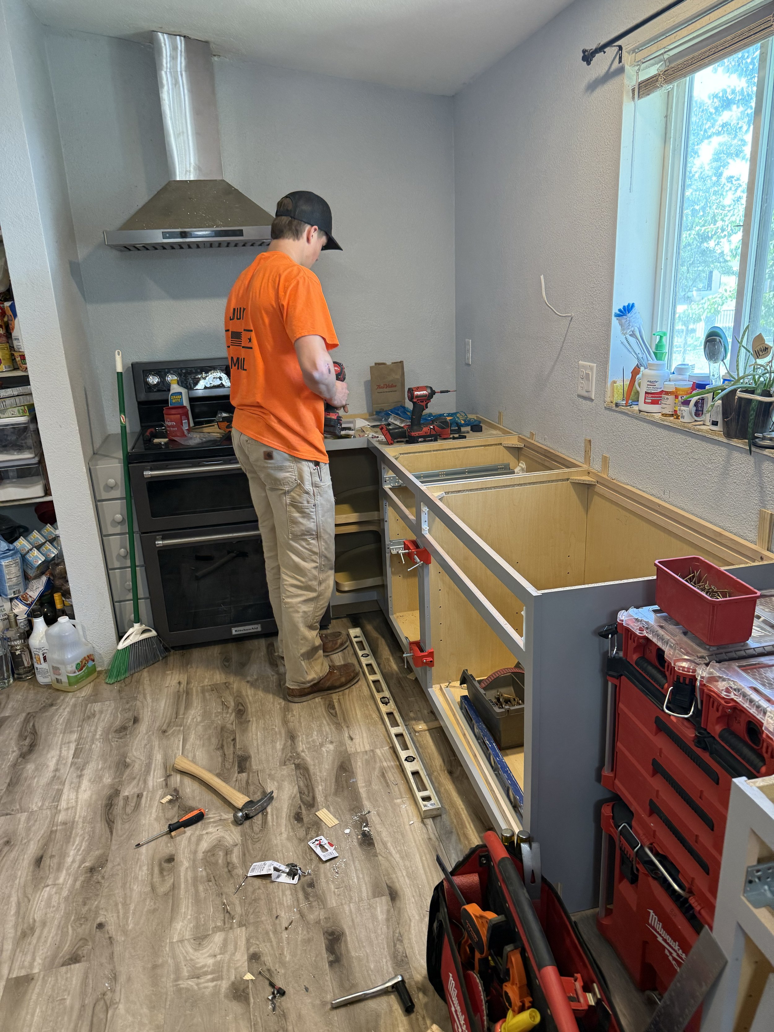 A man installing a kitchen cabinet frame with tools on the floor and a window on the right side.