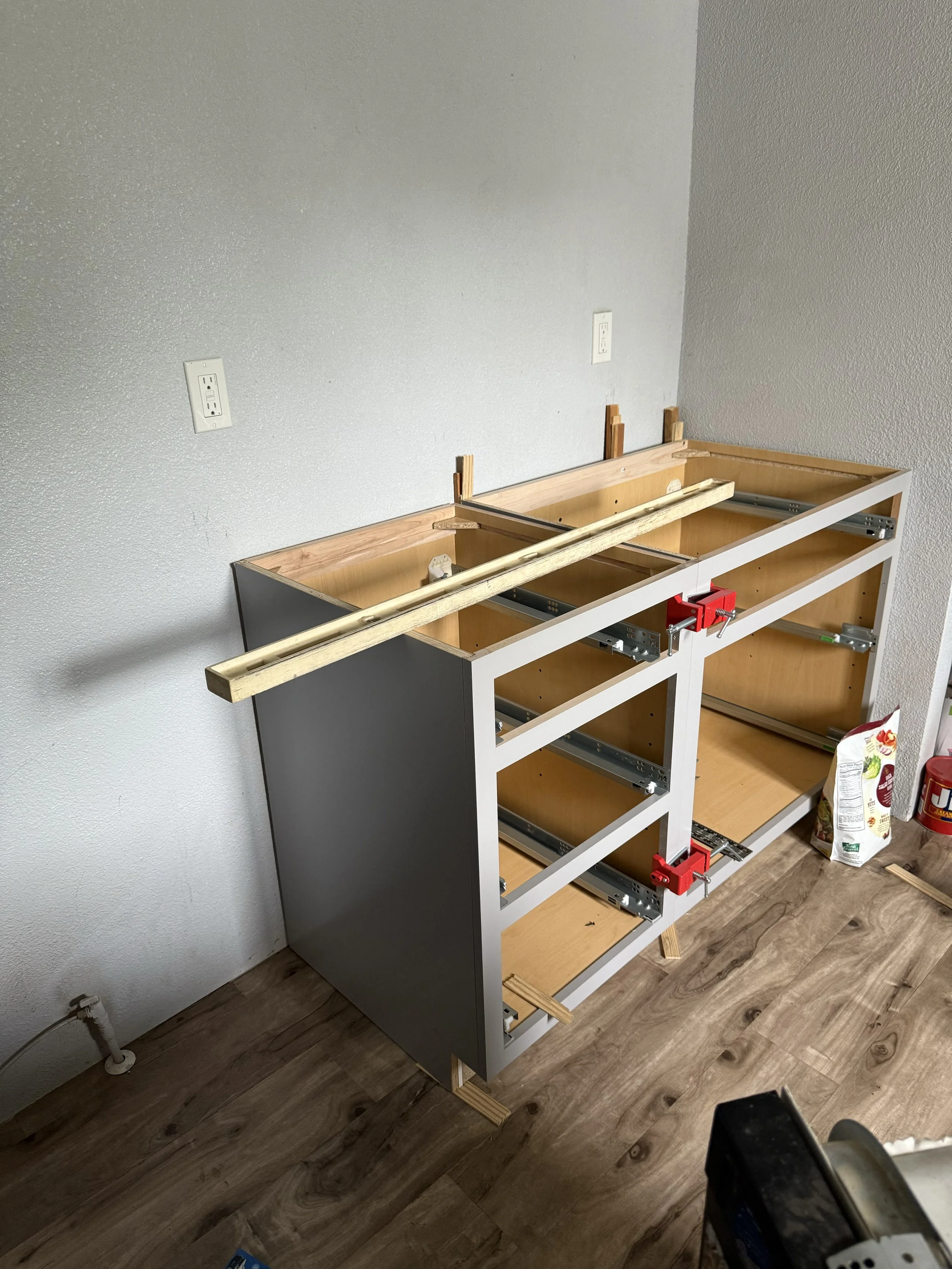 Kitchen cabinet under construction with wooden frame, being assembled against a wall, on a wooden floor, with tools and materials nearby.