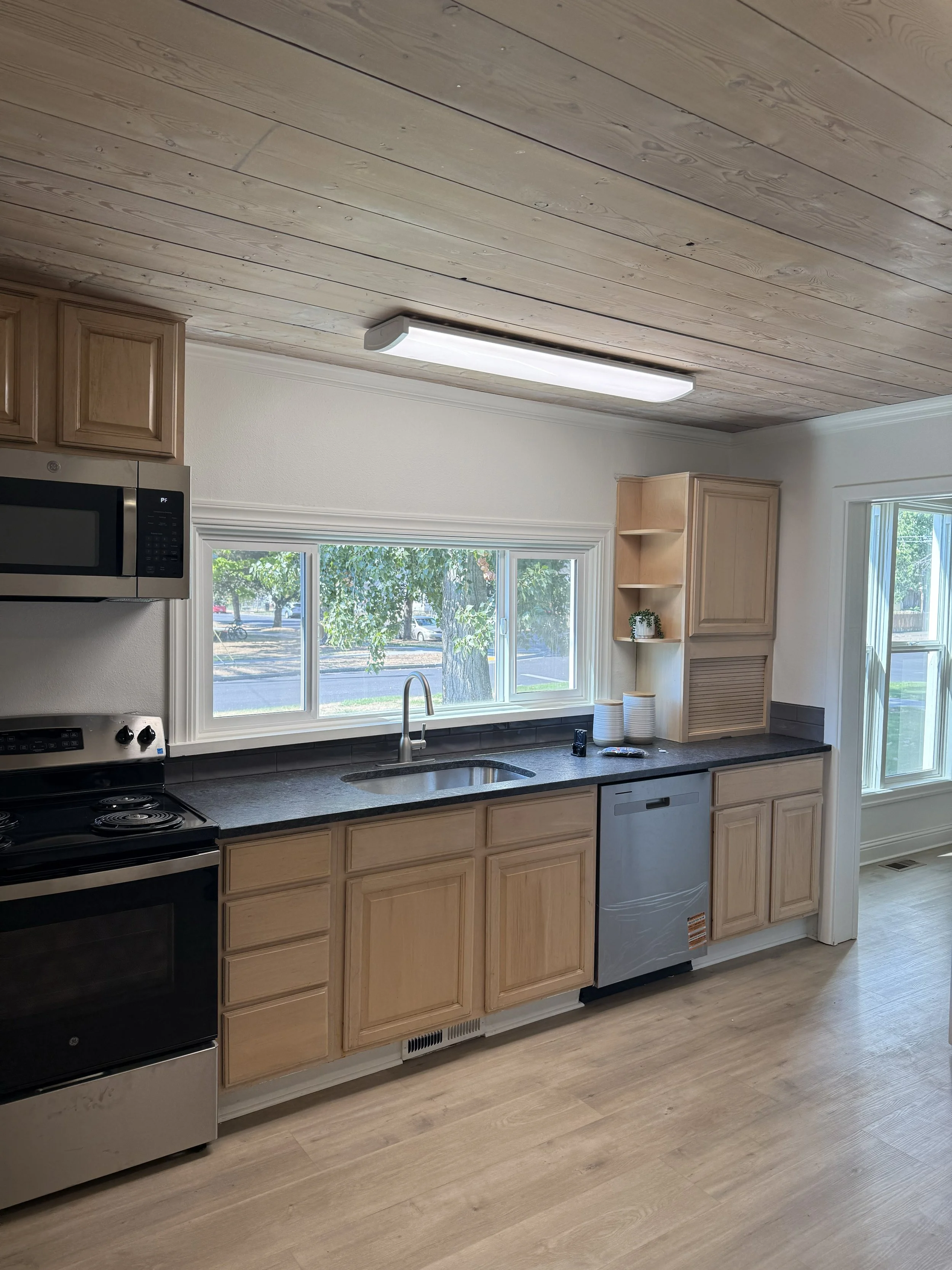 Kitchen with wood cabinets, black countertop, stainless steel appliances, and a large window over the sink.
