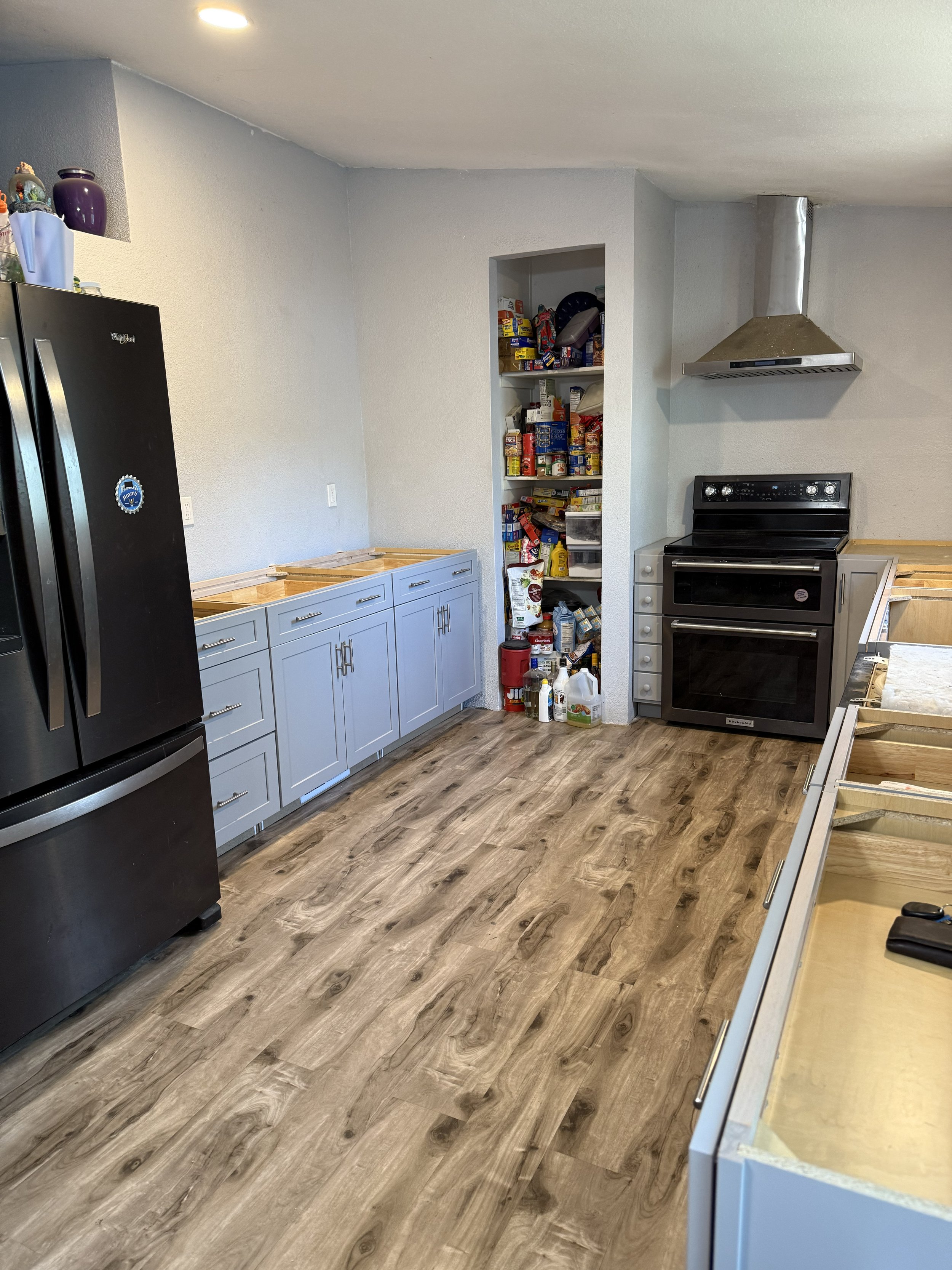 Kitchen with black refrigerator, light blue cabinets, a black oven, and a pantry filled with canned goods and supplies. Wooden flooring.