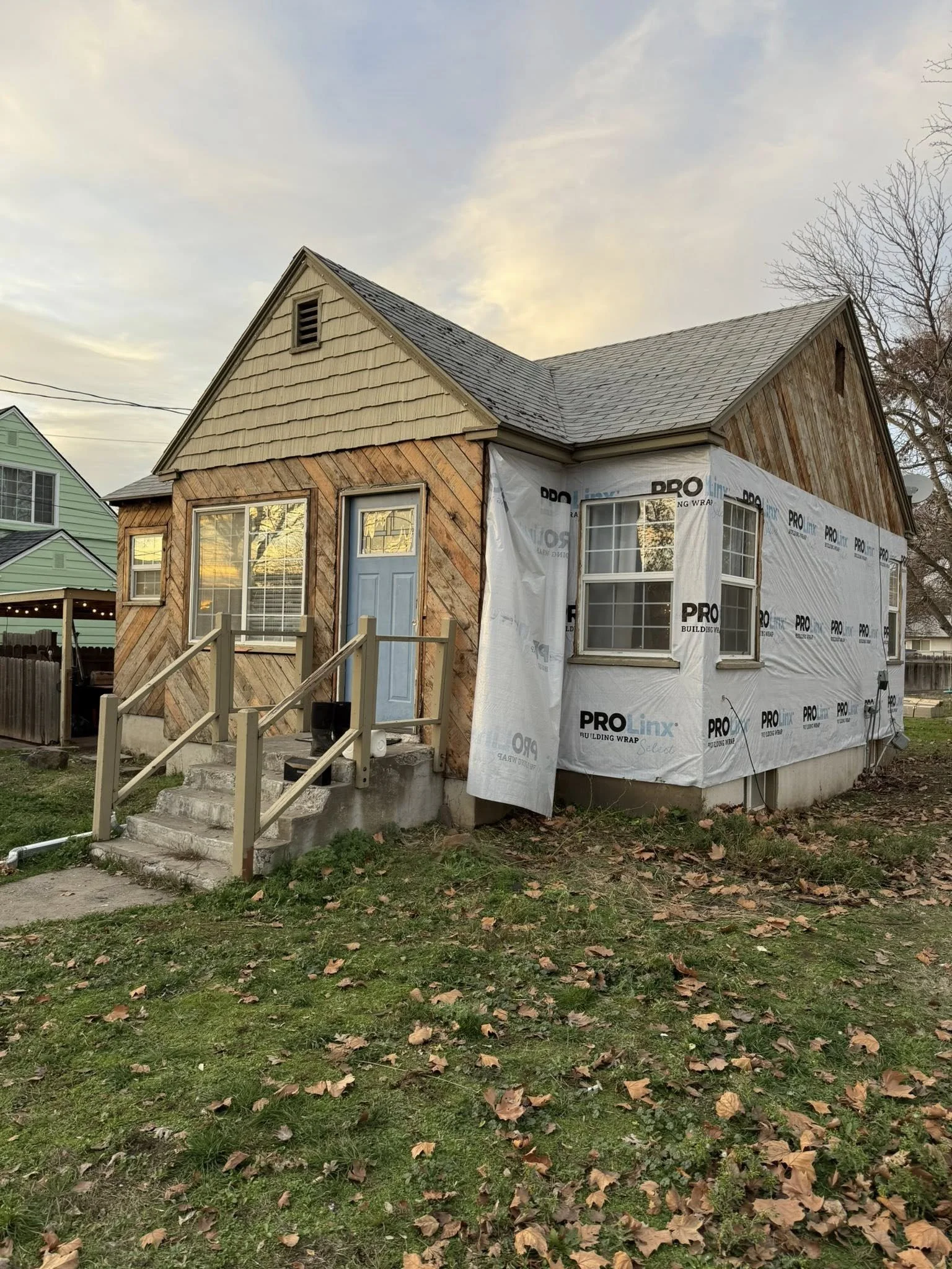 Two-story house under renovation with new siding being installed on one side, featuring a blue front door, concrete stairs with wooden railing, and a grassy yard with fallen leaves.