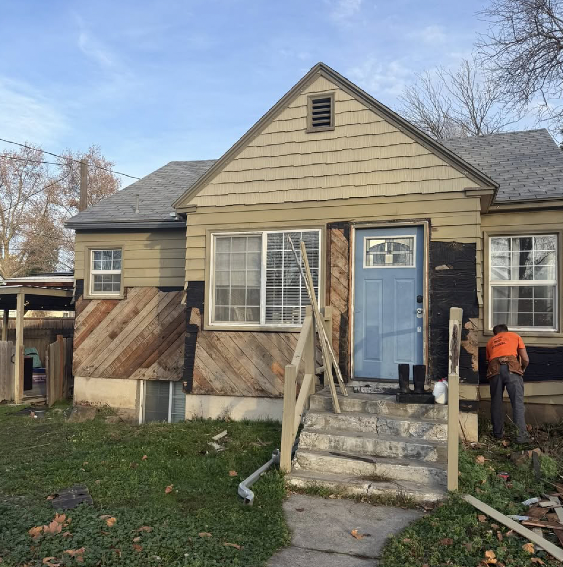 A house undergoing exterior renovation with siding removal; a contractor is working near the front entrance, and new wood is being installed around the door and windows.