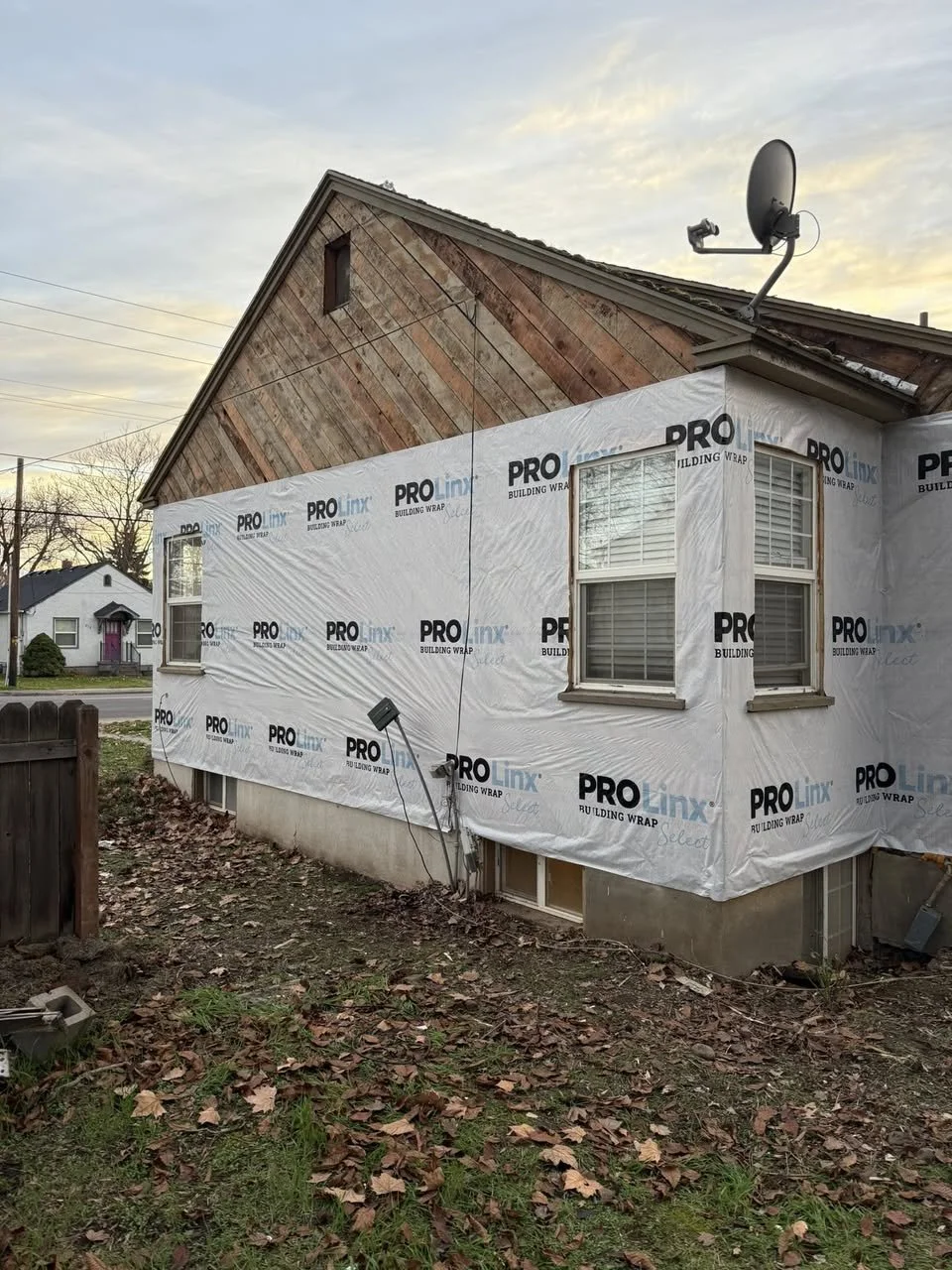 House undergoing renovation with new siding being installed, visible wrapping, two windows, satellite dish, and outdoor wiring, with fallen leaves on the ground.