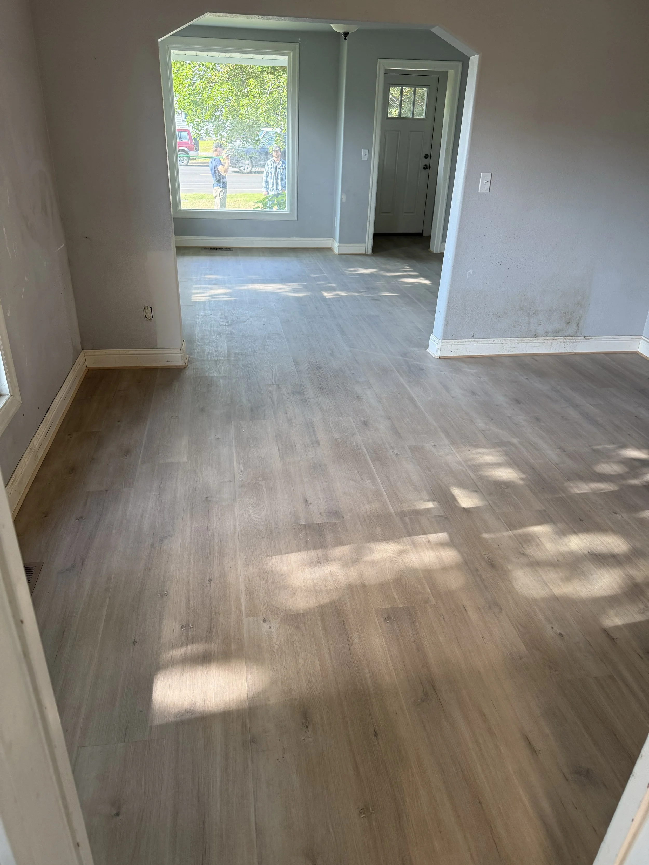 Empty room with light wood flooring, light gray walls, large front window, and doorway leading to an exterior view with trees and two people outside.