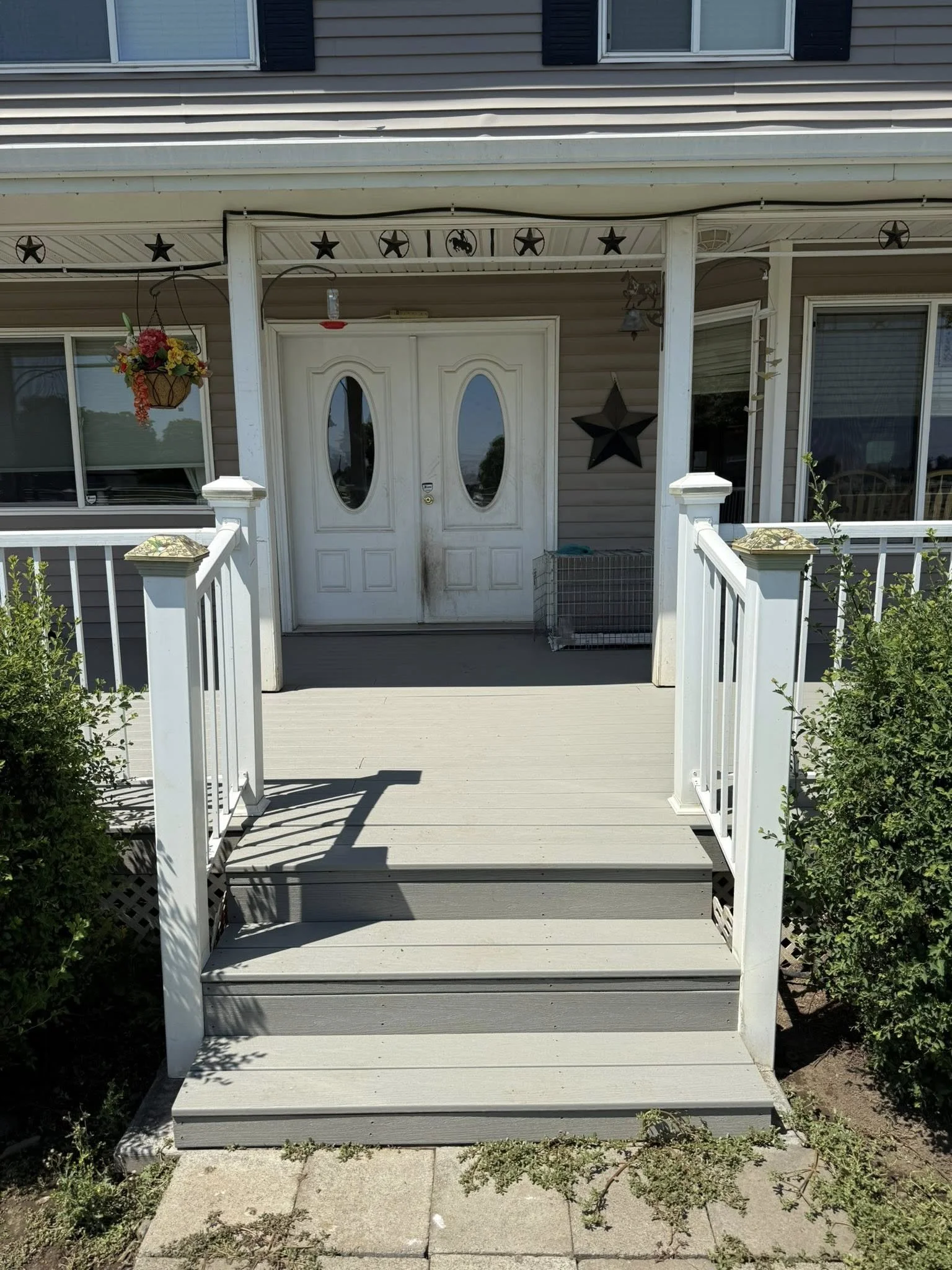 Front view of a house with a small porch, white double front doors with oval windows, hanging flower basket on the left, decorative stars on the wall, and bushes on both sides.