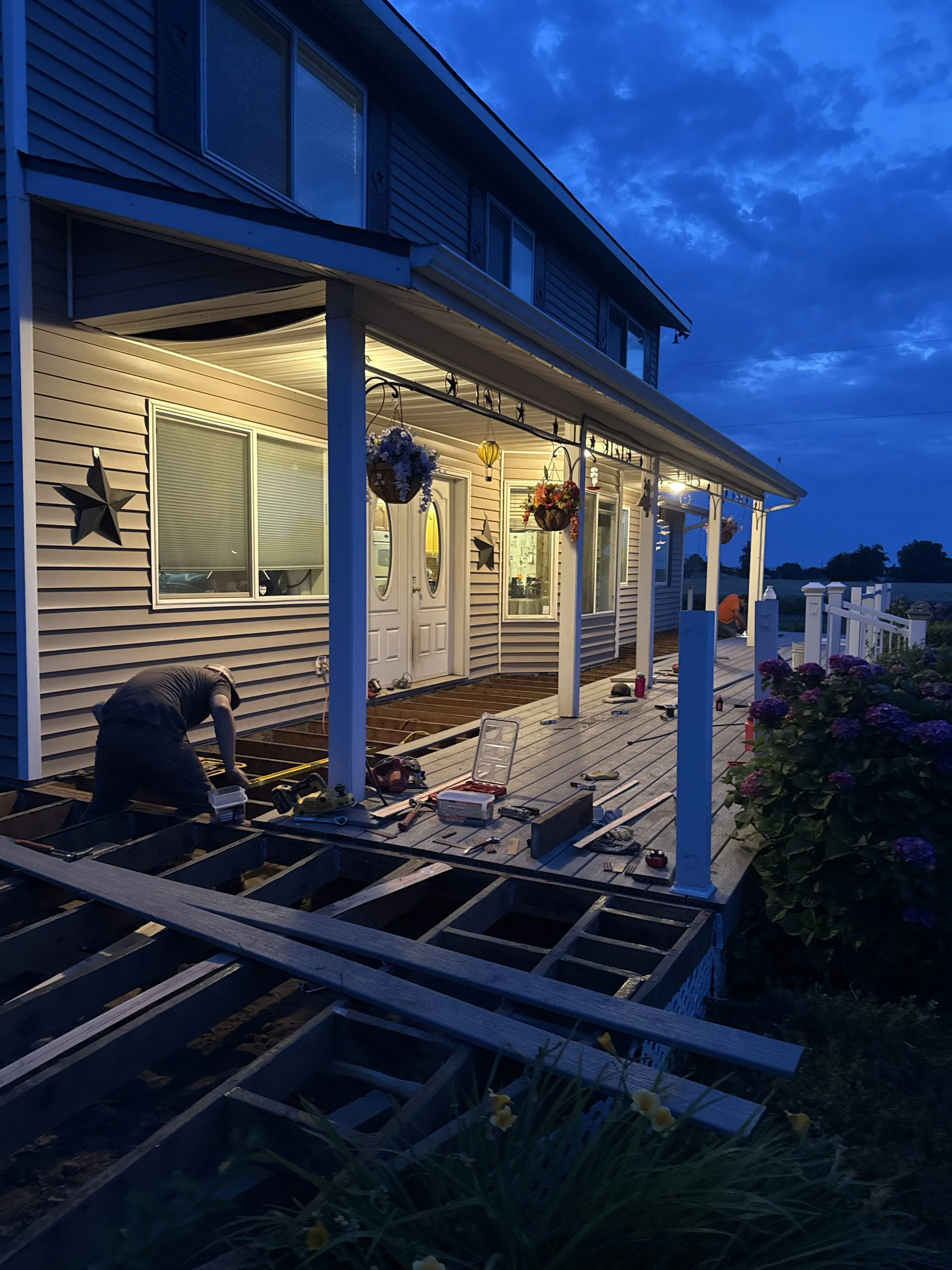 People working on decking construction on the porch of a house during evening, with tools and materials scattered around, and hanging flower baskets and star decorations on the house exterior.