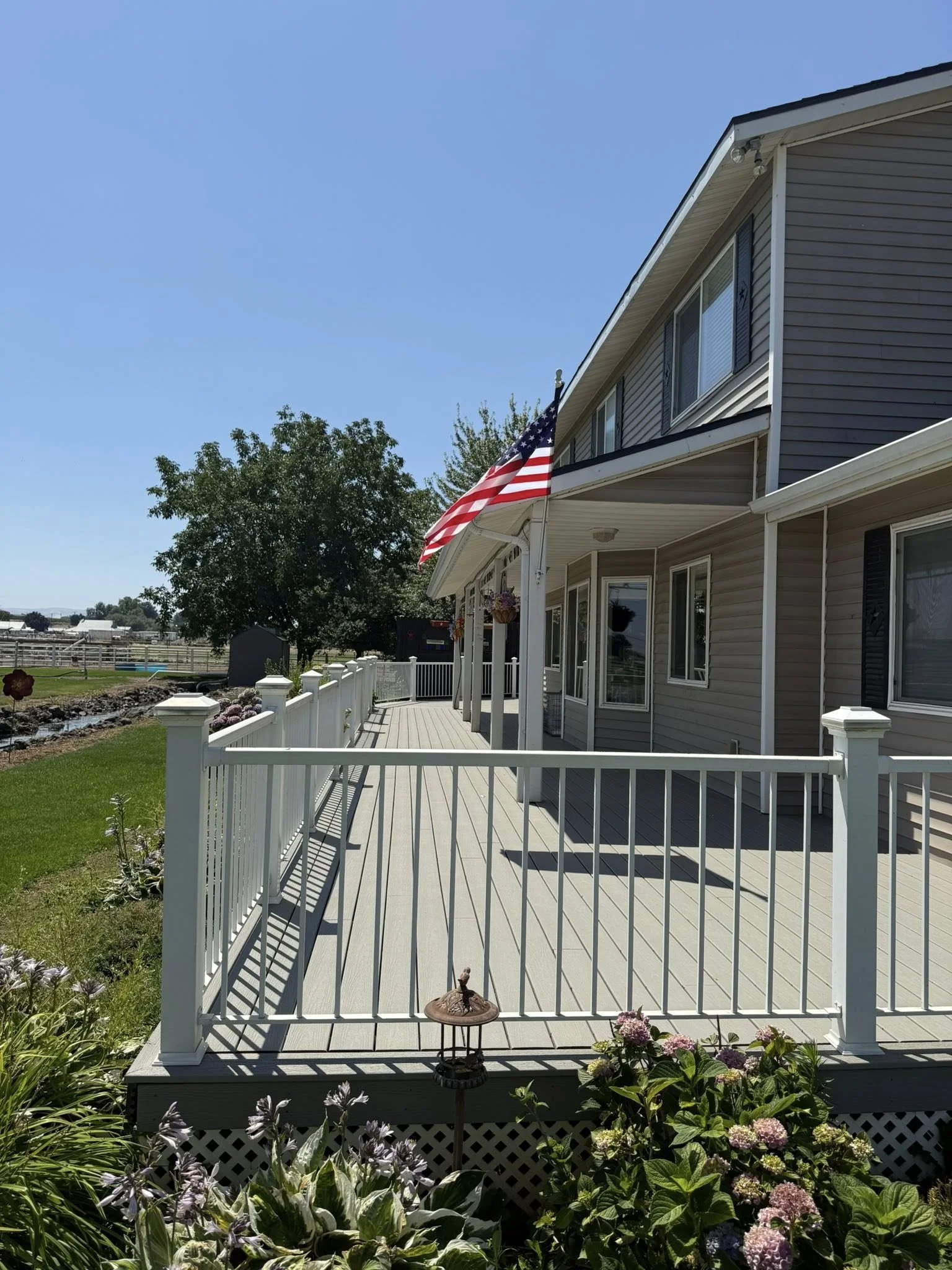 American flag hanging on the porch of a house with beige siding, white railing, and flower baskets, under a clear blue sky.