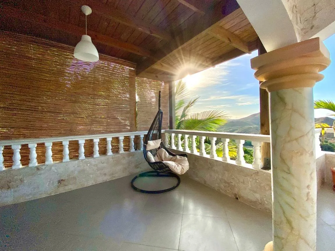 Terrasse ensoleillée avec un fauteuil suspendu, colonne en pierre, vue sur des collines verdoyantes, palmier, et un plafond en bois avec une lampe suspendue.