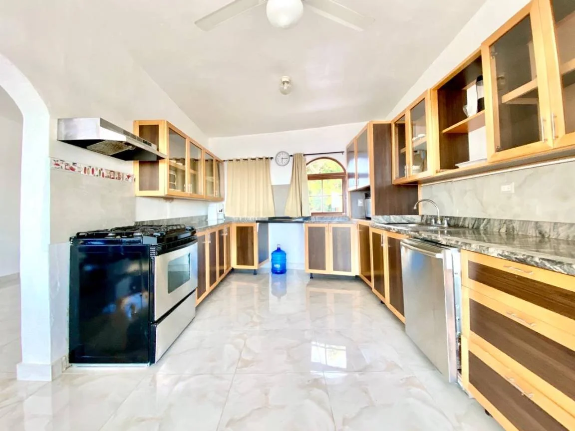 Cuisine spacieuse et lumineuse avec des armoires en bois, un four noir, un évier double et un grand plan de travail en marbre. Sol en carrelage et rideaux beige.