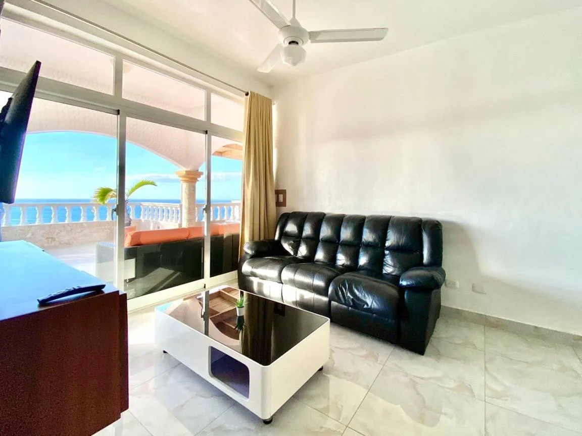 Un salon moderne avec un canapé en cuir noir, une table basse blanche et noire, un ventilateur de plafond, une télévision et une baie vitrée donnant sur une terrasse avec vue sur la mer.