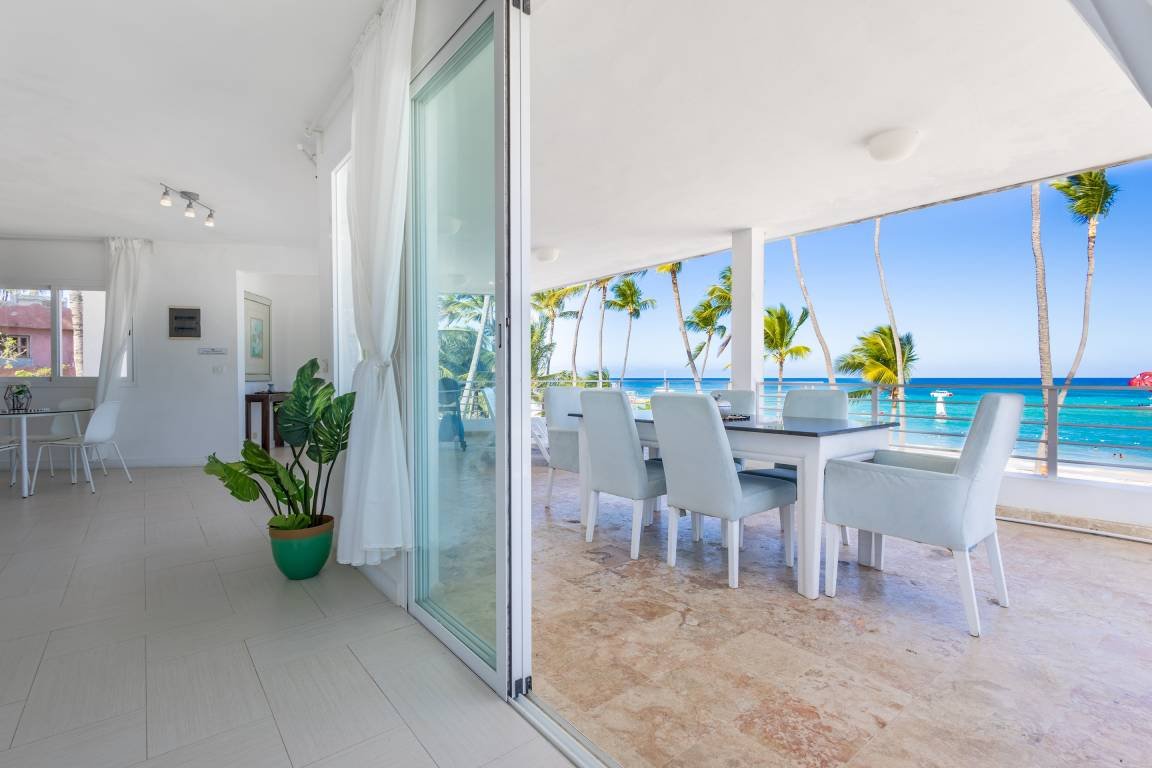 Intérieur lumineux avec table, chaises et plantes, vue sur terrasse avec table extérieure et mer en arrière-plan.