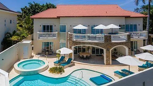 Grande villa avec piscine, jacuzzi, chaises longues et parasols, entourée de végétation tropicale.