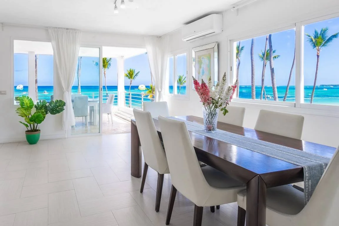 Salle à manger lumineuse avec vue sur la mer, des chaises blanches autour d'une table en bois foncé, vase avec fleurs, plantes en pot, grandes fenêtres et portes vitrées donnant sur un balcon avec palmiers et mer turquoise.
