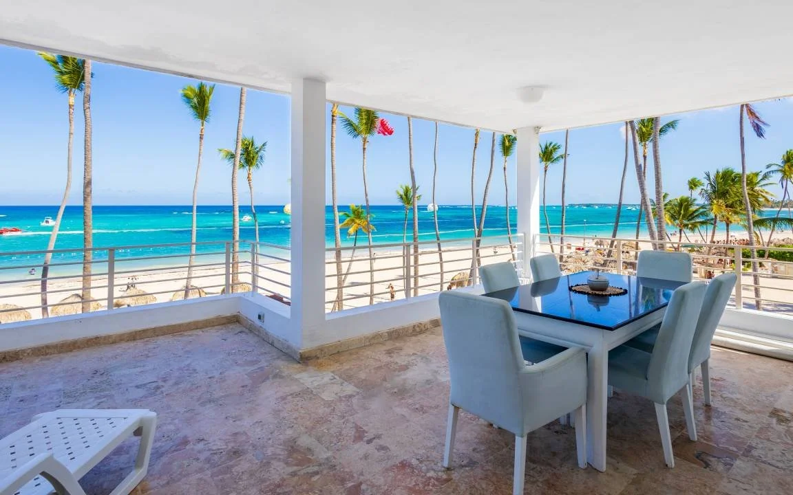 Terrasse avec mobilier d'extérieur surplombant une plage de sable et l'océan turquoise avec des palmiers.
