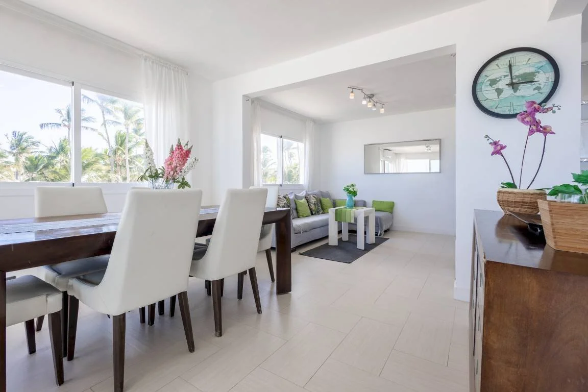 Intérieur lumineux avec salle à manger et salon, table en bois, chaises blanches, canapé gris, coussins verts, décoration florale, horloge murale, grandes fenêtres avec vue sur des palmiers.